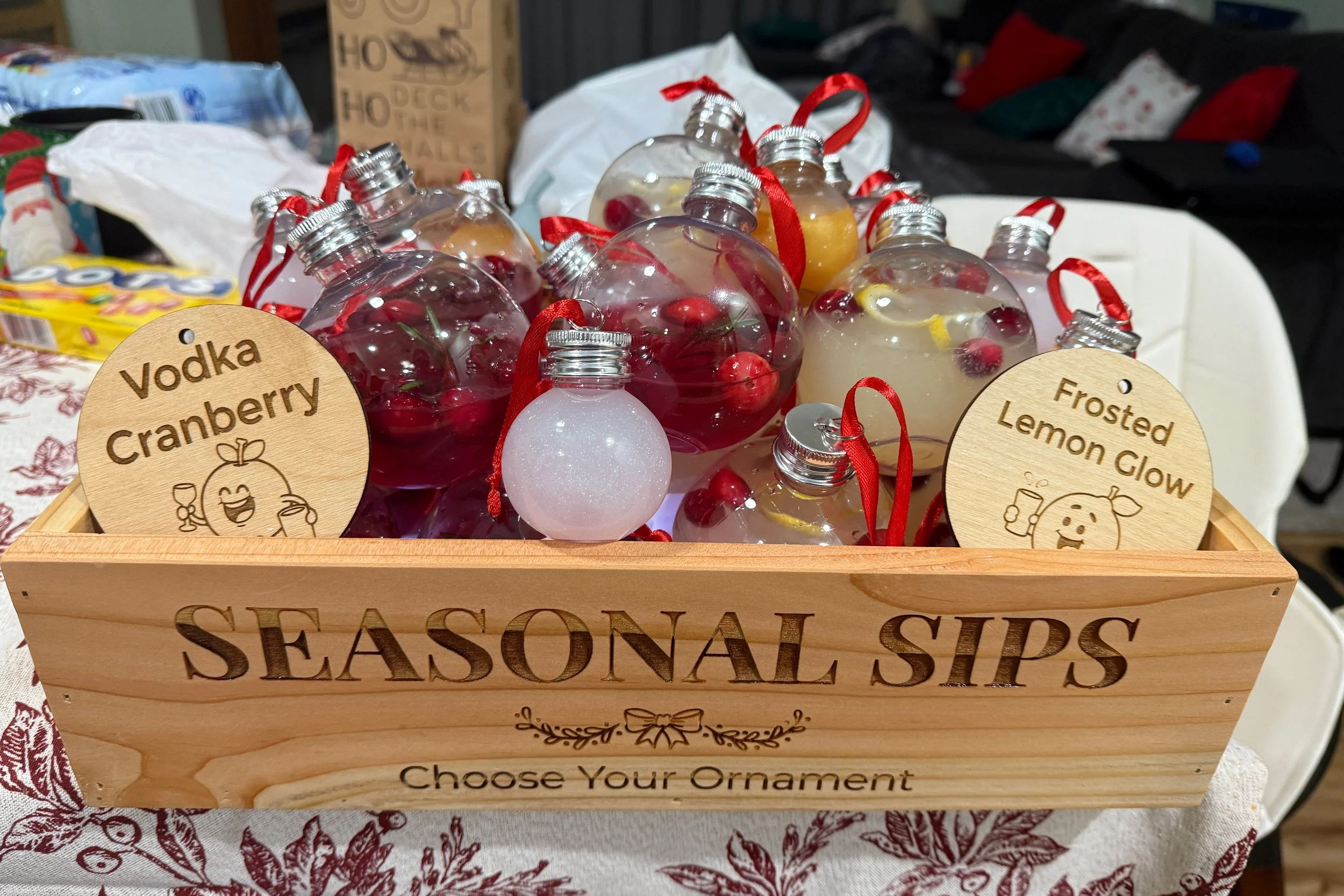 A wooden box with the words "Seasonal sips choose your ornament" engraved on the front, filled with ornaments with juice and fruits and two wooden engraved Christmas ornaments with sayings and retro styled cartoons