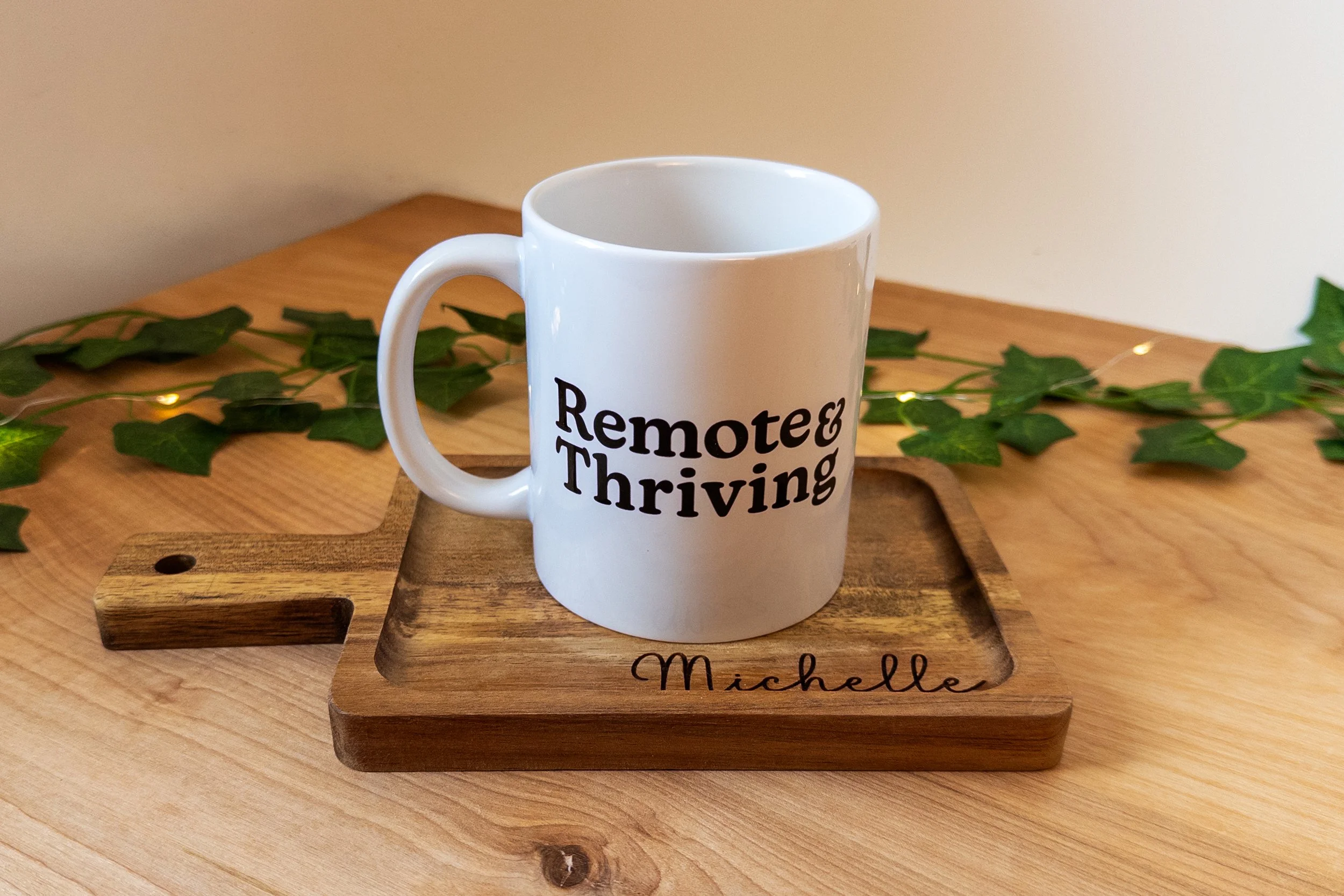 11oz coffee mug that says "Remote & Thriving" on a personalized sized cutting board with the name "Michelle" engraved on it, sitting on a wooden table with ivy behind it. 