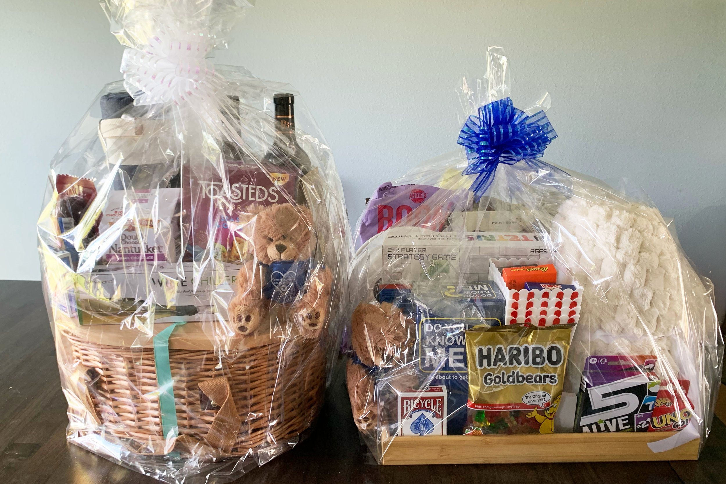 Two big gift baskets wrapped in cellophane sitting on a table. One a picnic theme, the other a game night theme. 