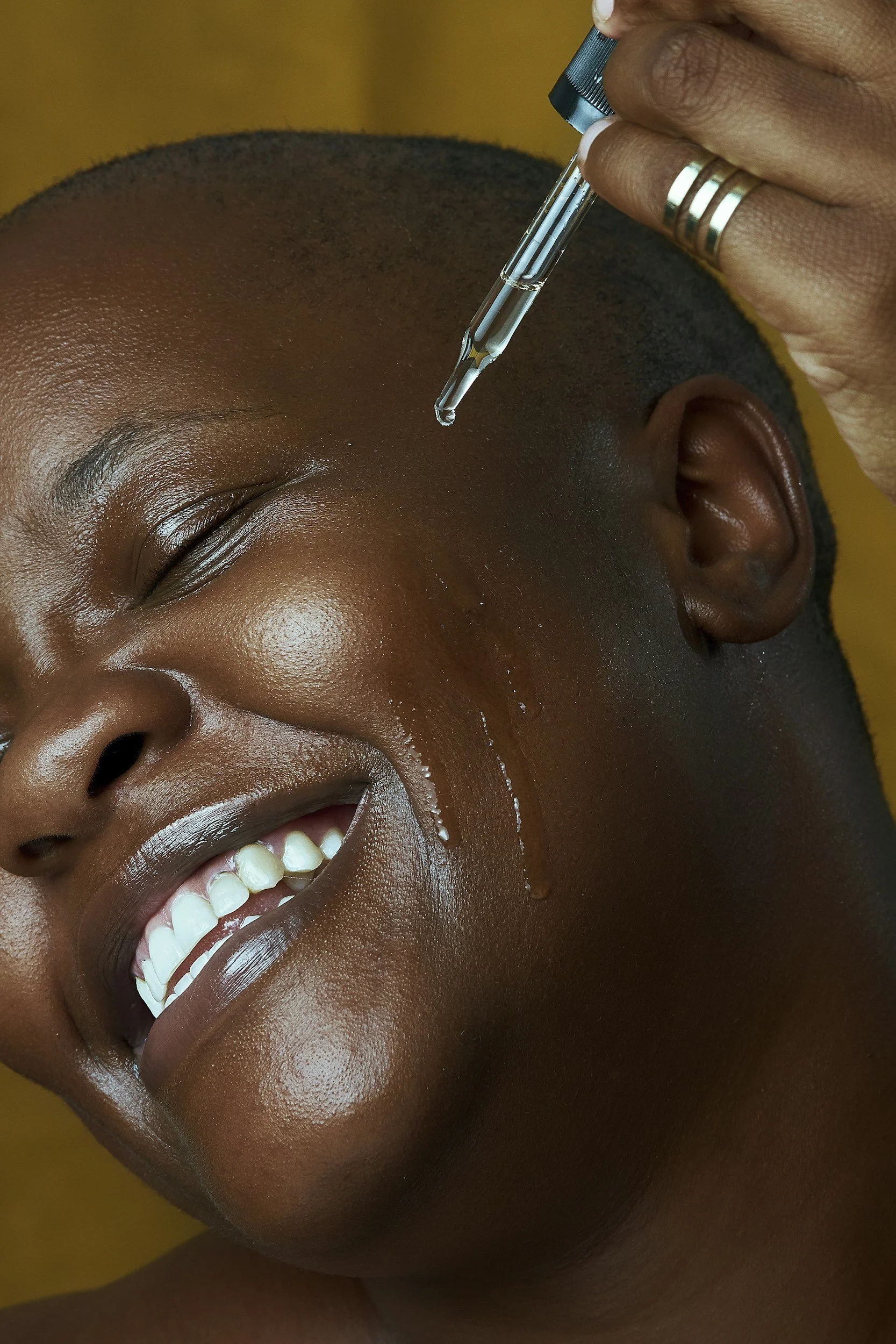 Person receiving an eyebrow treatment with a dropper, smiling with closed eyes and showing white teeth.