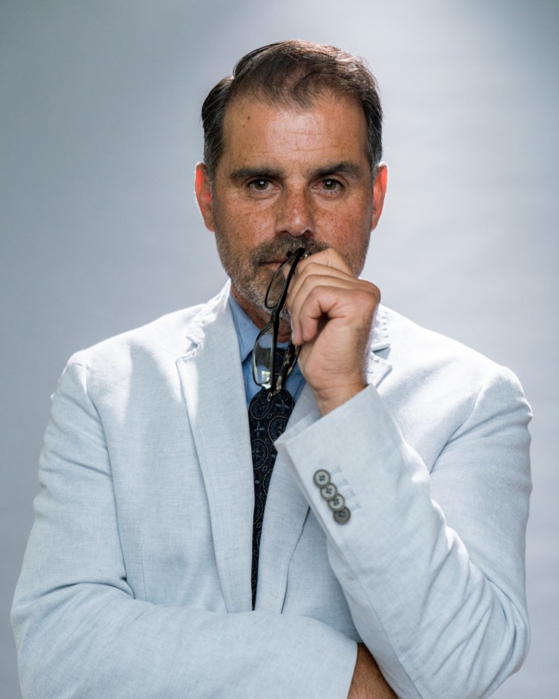 A man with short dark hair, beard, and mustache wearing a white blazer holding glasses near his mouth, looking into the camera with a serious expression, against a gray background.