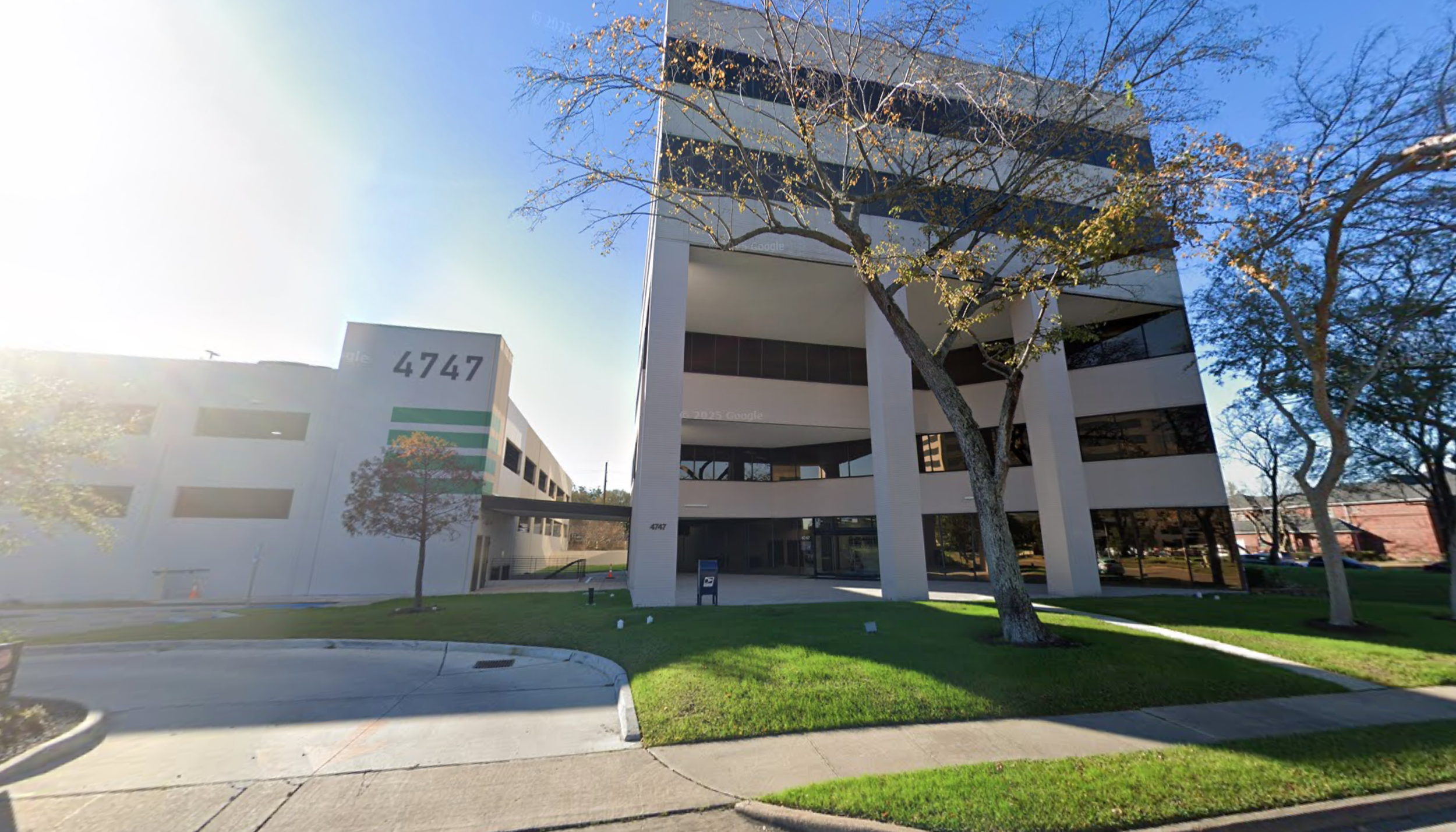 Modern office building with white exterior, black balconies, and the number 4747 on the side. In front, there are trees with some leaves changing color, green grass, sidewalk, and street.