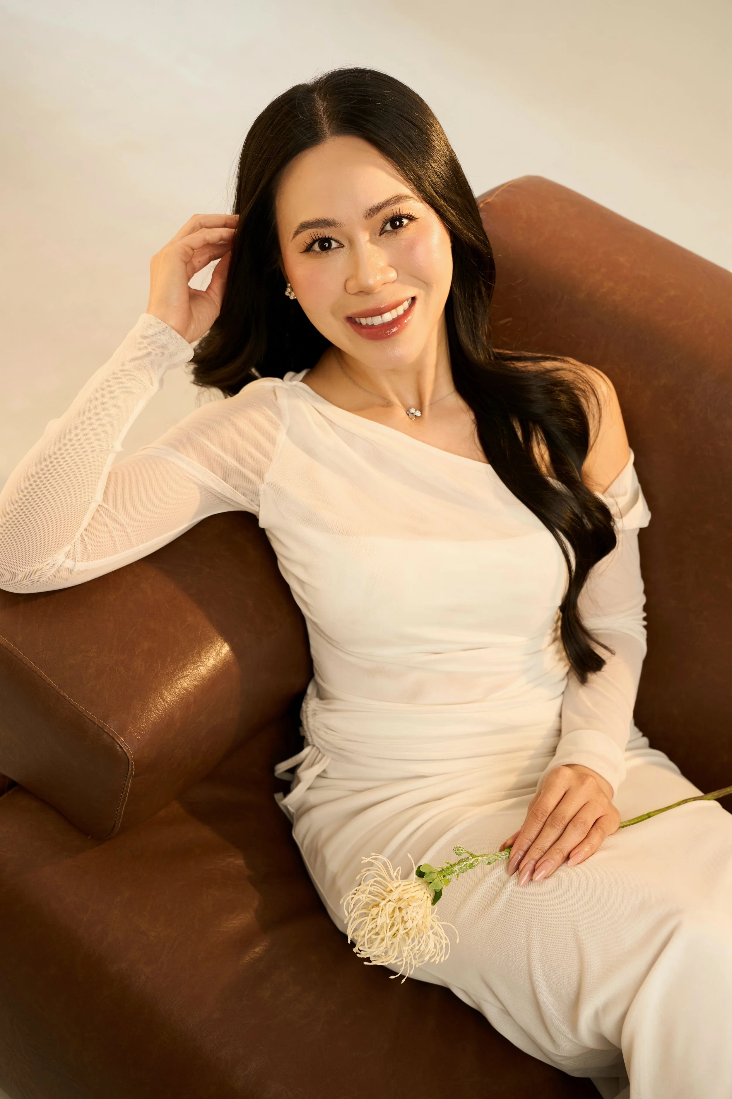 A woman with long black hair and a white dress sitting on a brown leather couch, holding a white flower, smiling at the camera.