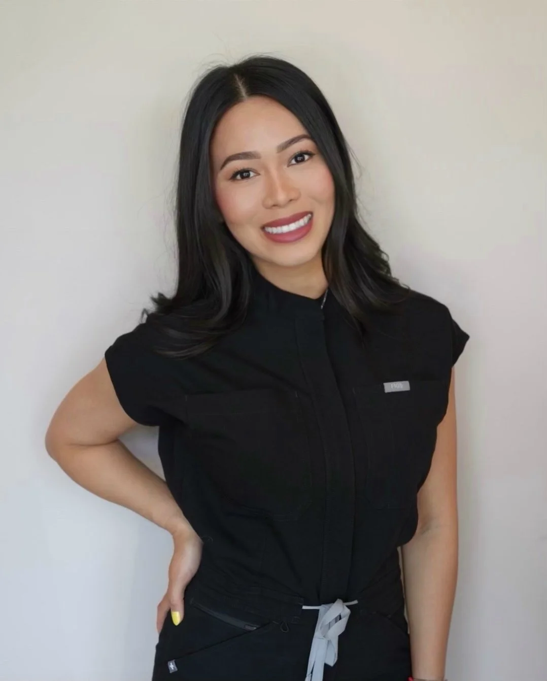Young woman with black hair smiling, wearing a black nurse uniform, posing with one hand on her hip against a plain background.