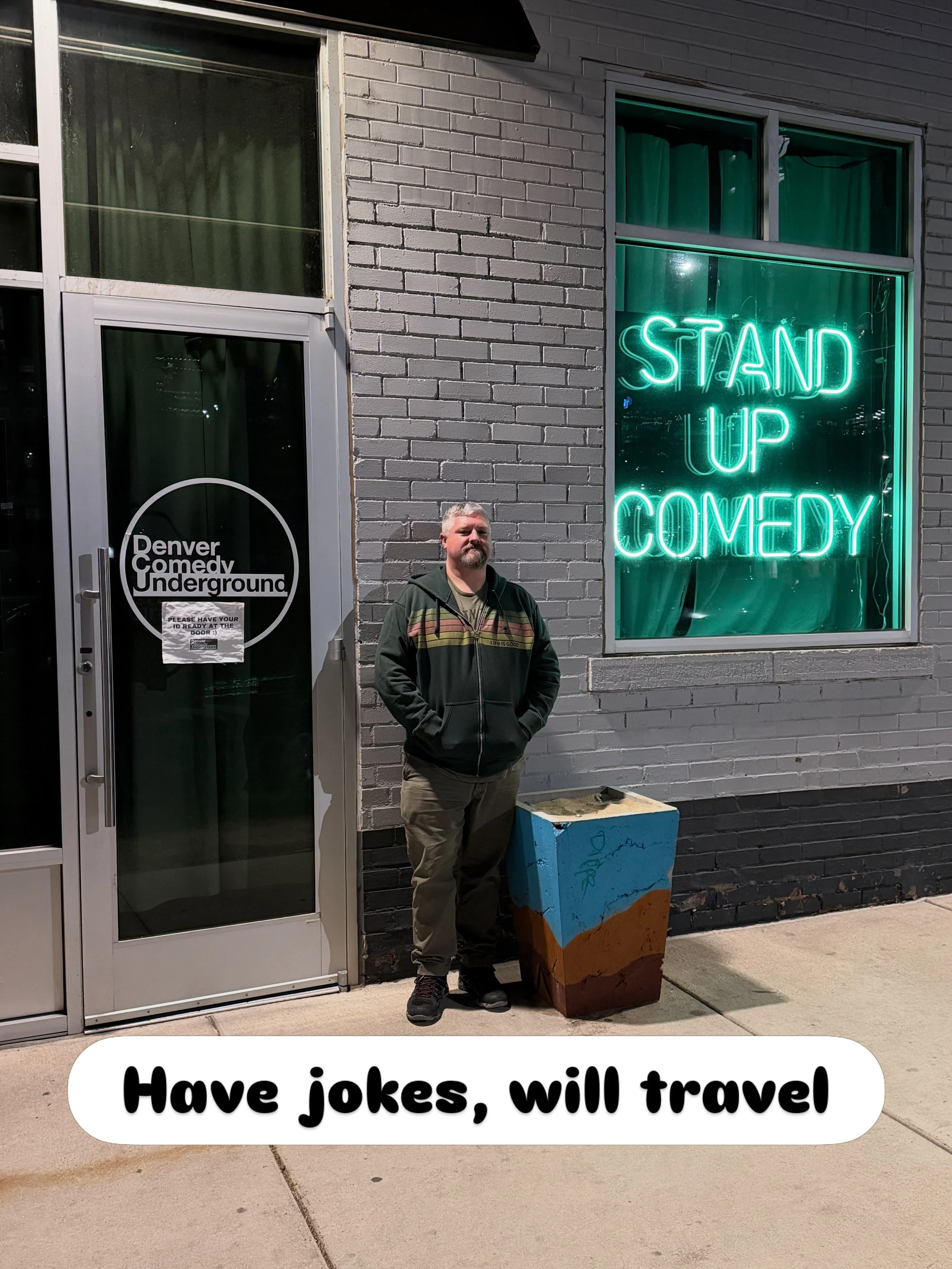A man standing outside a building with a sign reading "STAND UP COMEDY" in neon lights. There is a door with a sign that says "Denver Comedy Underground" and a painted flowerpot in front of him. A caption at the bottom says "Have jokes, will travel".