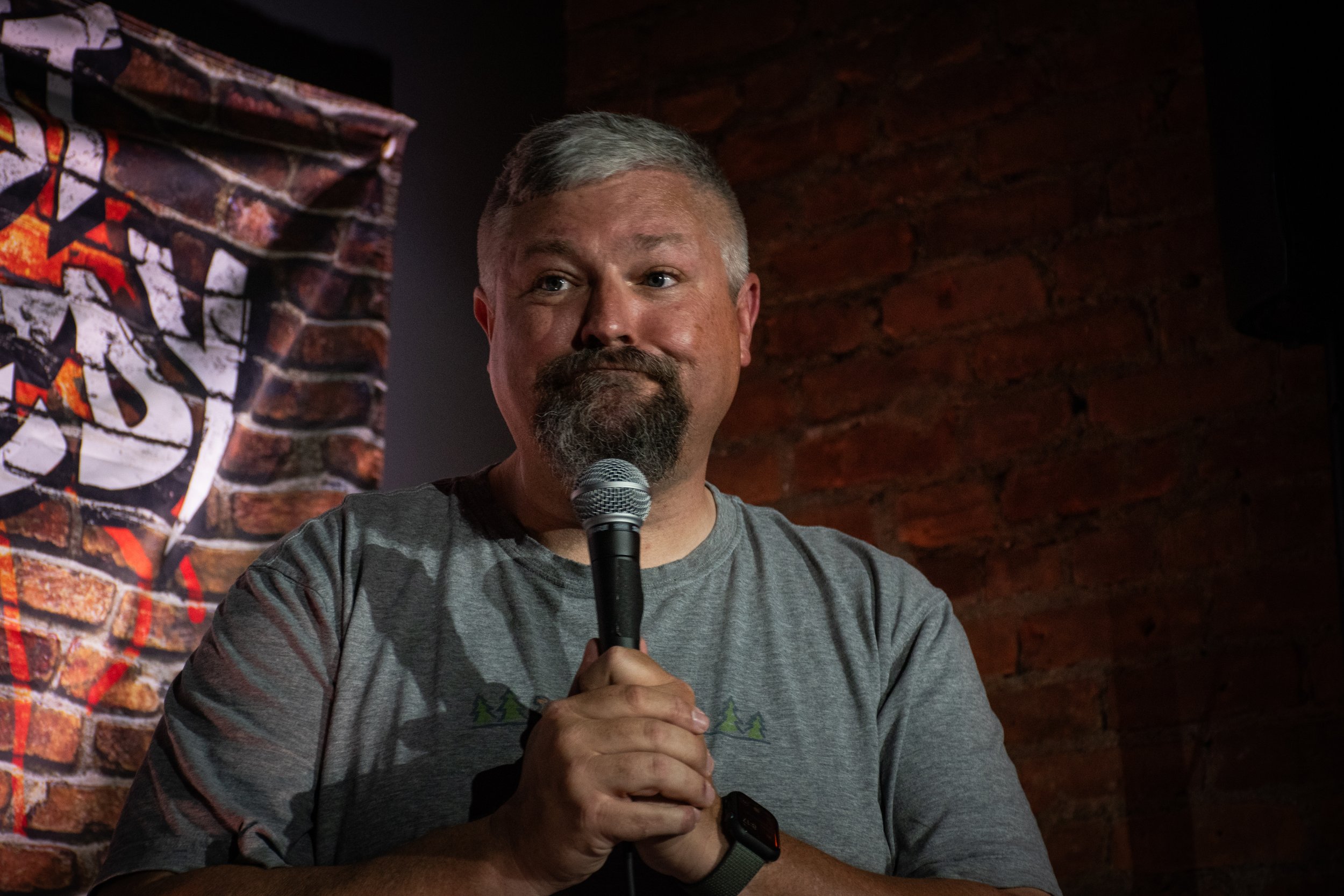 A man with gray hair, beard, and mustache, holding a microphone, standing in front of a brick wall with a graffiti-style poster partially visible.