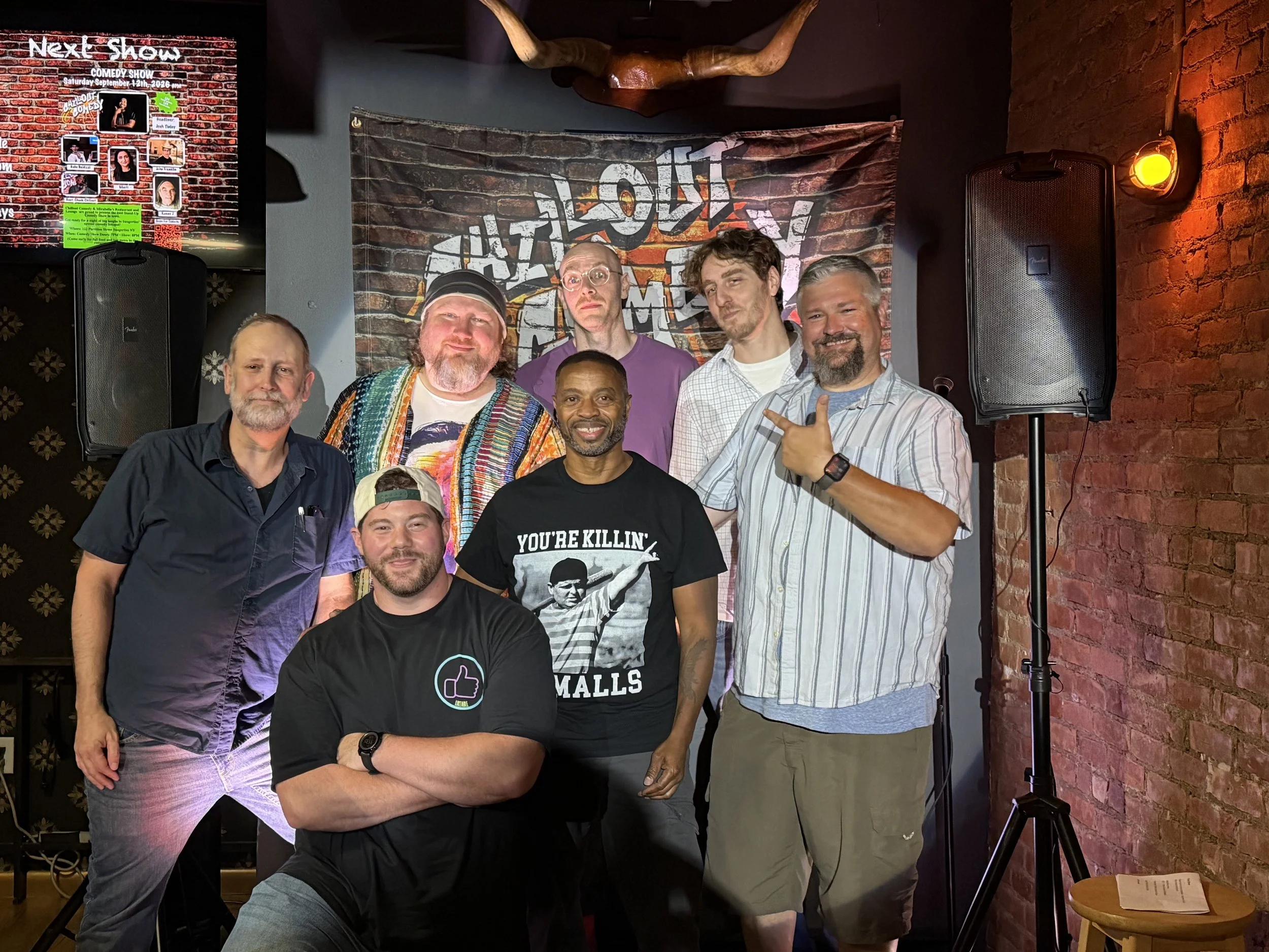 A group of seven men posing together indoors, with brick walls and stage equipment in the background. They are smiling and standing in front of a backdrop with graffiti-style writing that says 'COMEDY.' Some of the men have facial hair, and one man is wearing a black shirt with text and an image of a person holding a microphone.