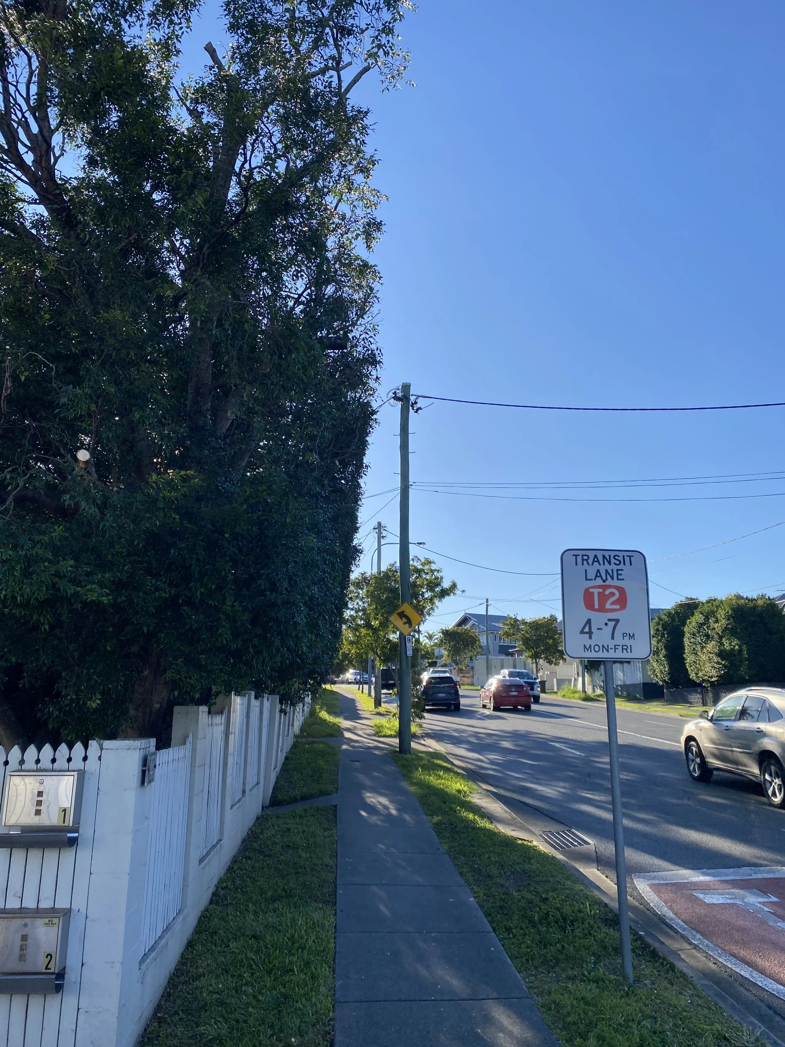 A suburban sidewalk scene during daytime with a white picket fence on the left, a row of mailbox boxes, trees, utility poles, cars parked along the street, and a transit lane sign indicating restrictions from 4 to 7 PM, Monday to Friday.