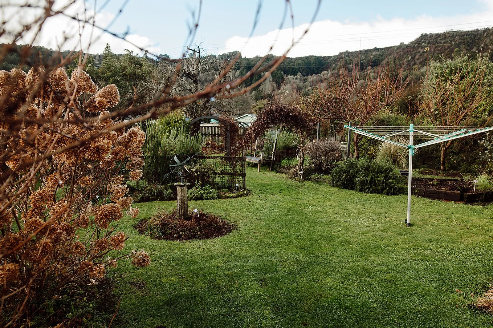 A lush backyard garden with green grass, flowering shrubs, trees, a birdbath, a bench, a garden arch, and a rotary clothesline, set against distant hills and a partly cloudy sky.