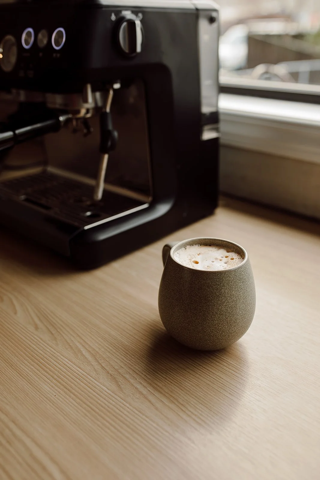 A mug of frothy coffee on a wooden table near an espresso machine.