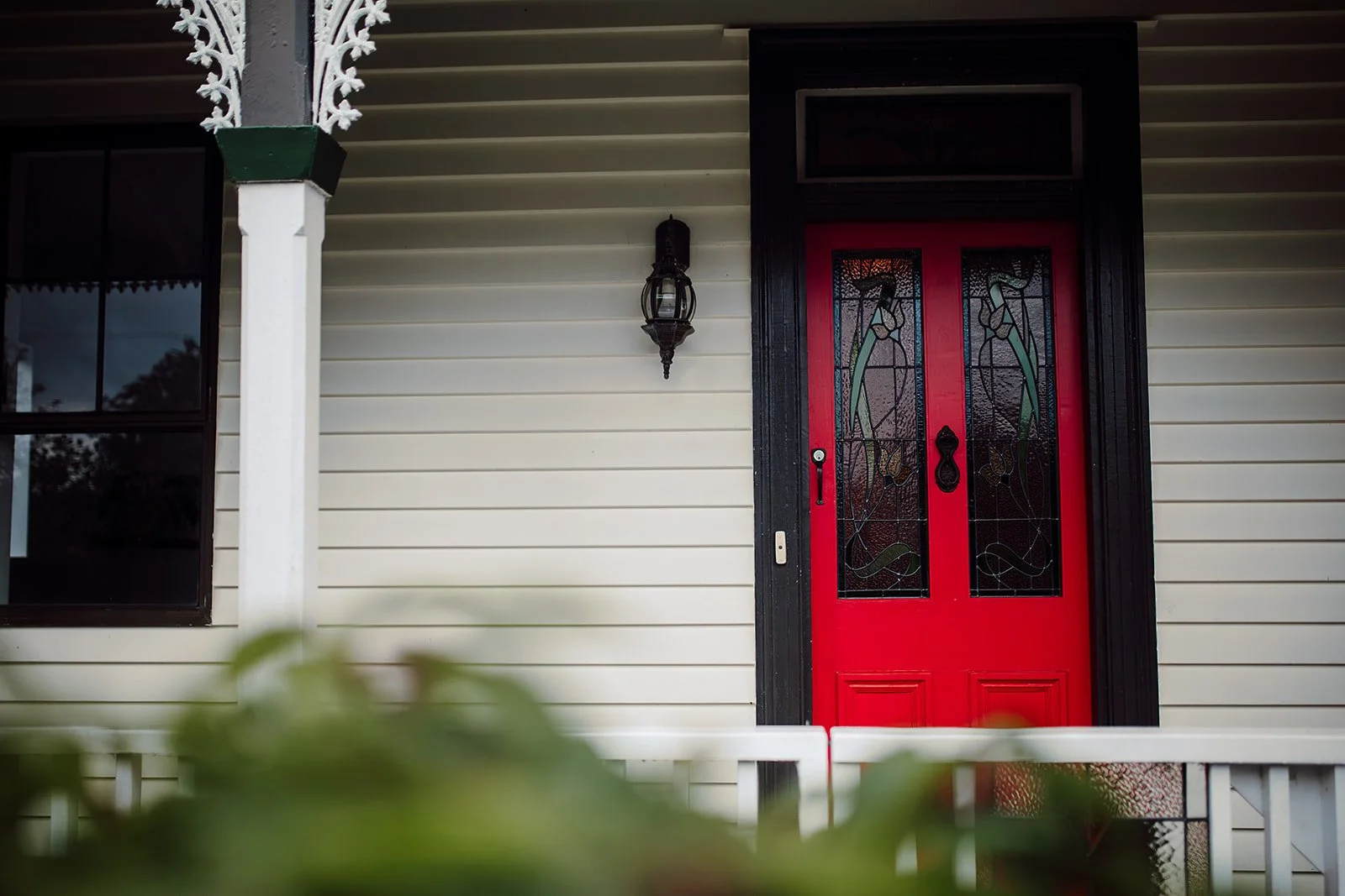 Red front door with stained glass panels decorated with flowers and leaves, black trim, and a decorative black door handle, on a house with beige horizontal siding and a black lantern-style light fixture beside it.