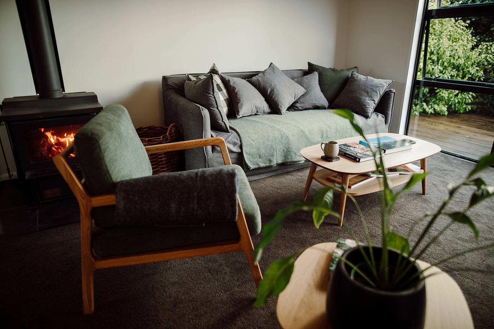 Cozy living room with a grey sofa, assortment of pillows, a small wooden coffee table with magazines and a coffee mug, a green armchair, a wood stove with a fire, and a glass door showing outdoor greenery.