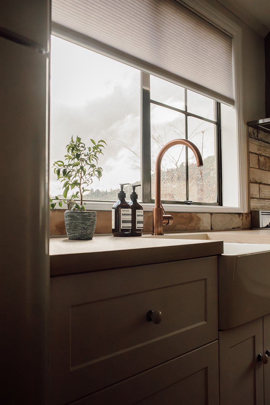 Kitchen sink with a brass faucet, a potted plant, and two soap dispensers on the countertop near a window with raindrops outside.
