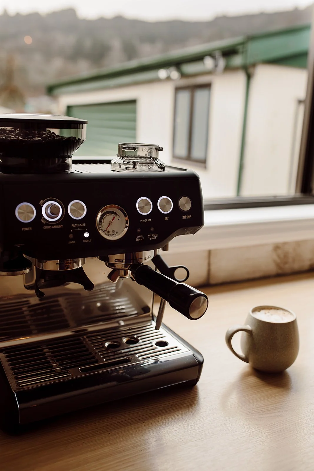 An espresso machine on a wooden countertop with a ceramic mug of coffee next to it, outside view of a building and mountains through a window.