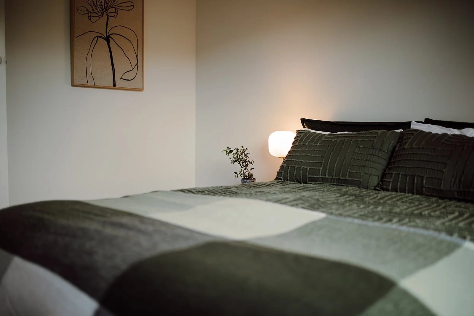 A neatly made bed with dark green and black patterned bedding in a well-lit bedroom, with a bedside pillow, a small plant, and a white lamp on the nightstand. There's a simple line drawing of a flower in a wooden frame hanging on the light-colored wa
