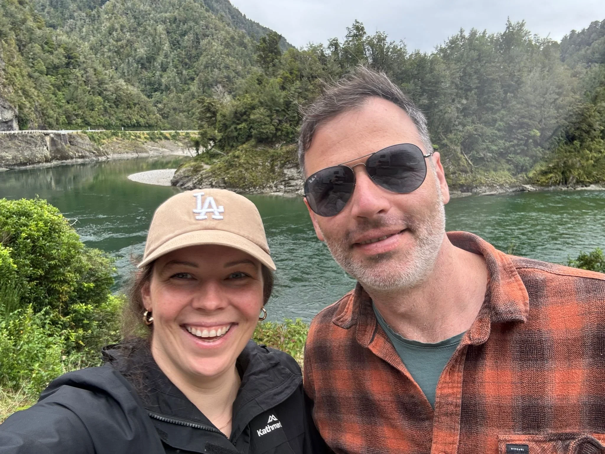Two people smiling outdoors near a river, with a forested mountain in the background. One person is wearing sunglasses and the other is wearing a beige cap with an 'LA' logo.