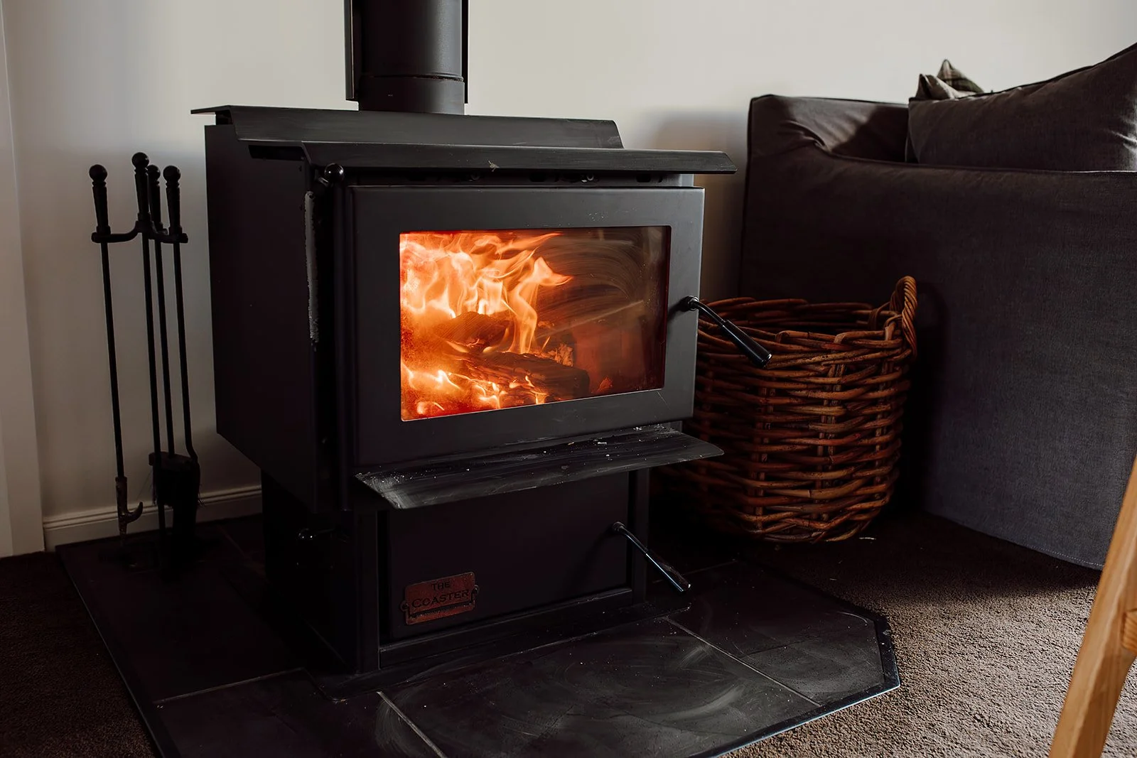 A black wood-burning stove with visible flames through a glass door, sitting on a black tiled hearth next to a wicker basket and a dark-colored sofa.
