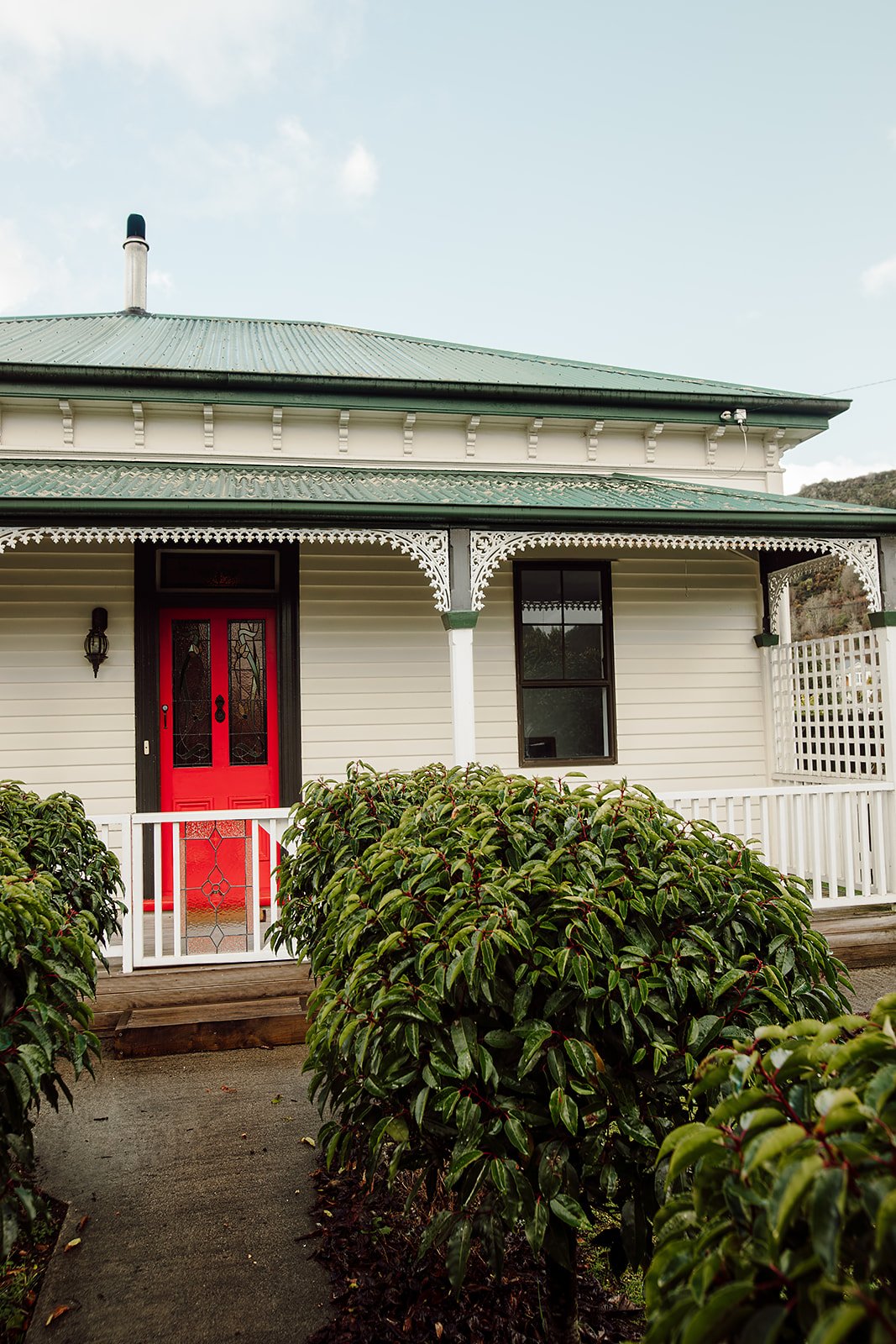 A house with a red front door, white siding, decorative trim, and a picket fence, surrounded by green bushes.