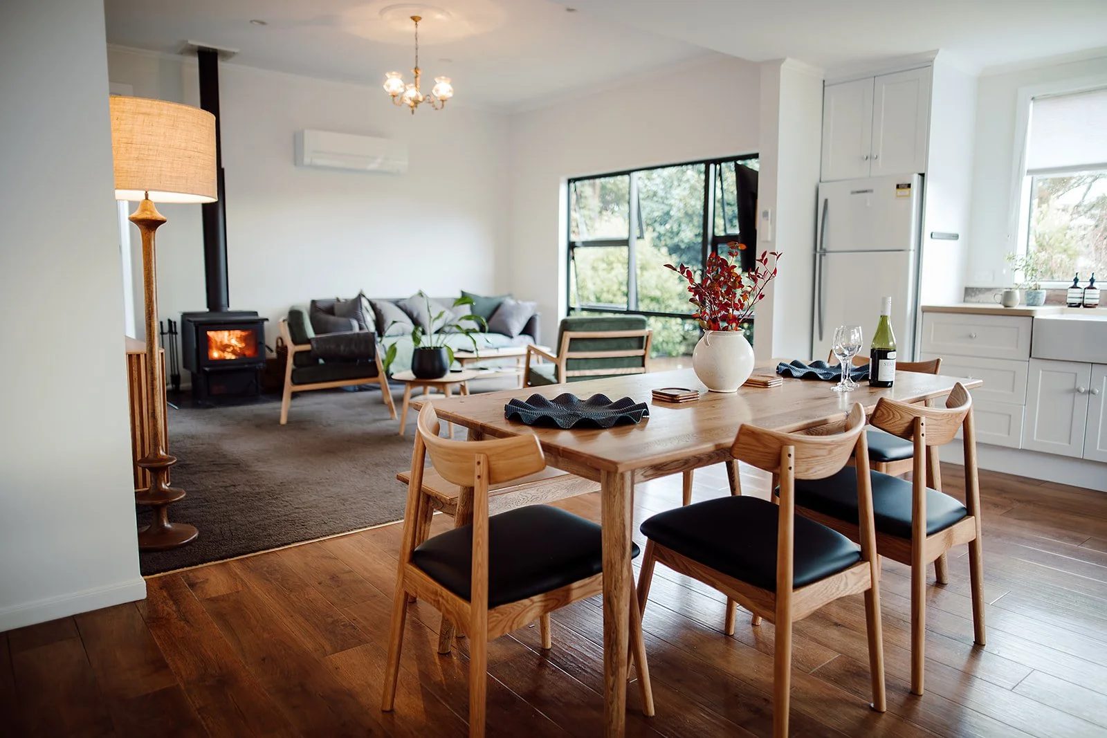 Open-concept living and dining area with a wooden dining table, six chairs, a white vase with red leaves, a bottle of wine, wine glasses, a black napkin, a white kitchen cabinet, a window with trees outside, a leather sofa, a coffee table, a wood sto