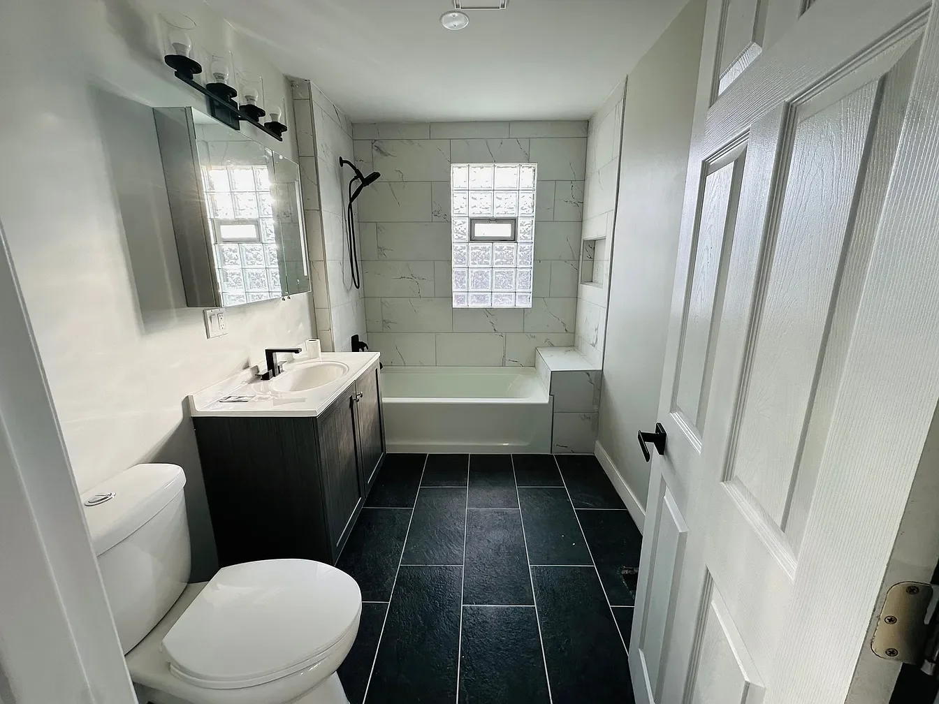 Modern bathroom with black floor tiles, white walls, a bathtub, a window with glass blocks, a black vanity with a white sink, a mirror, and a toilet.