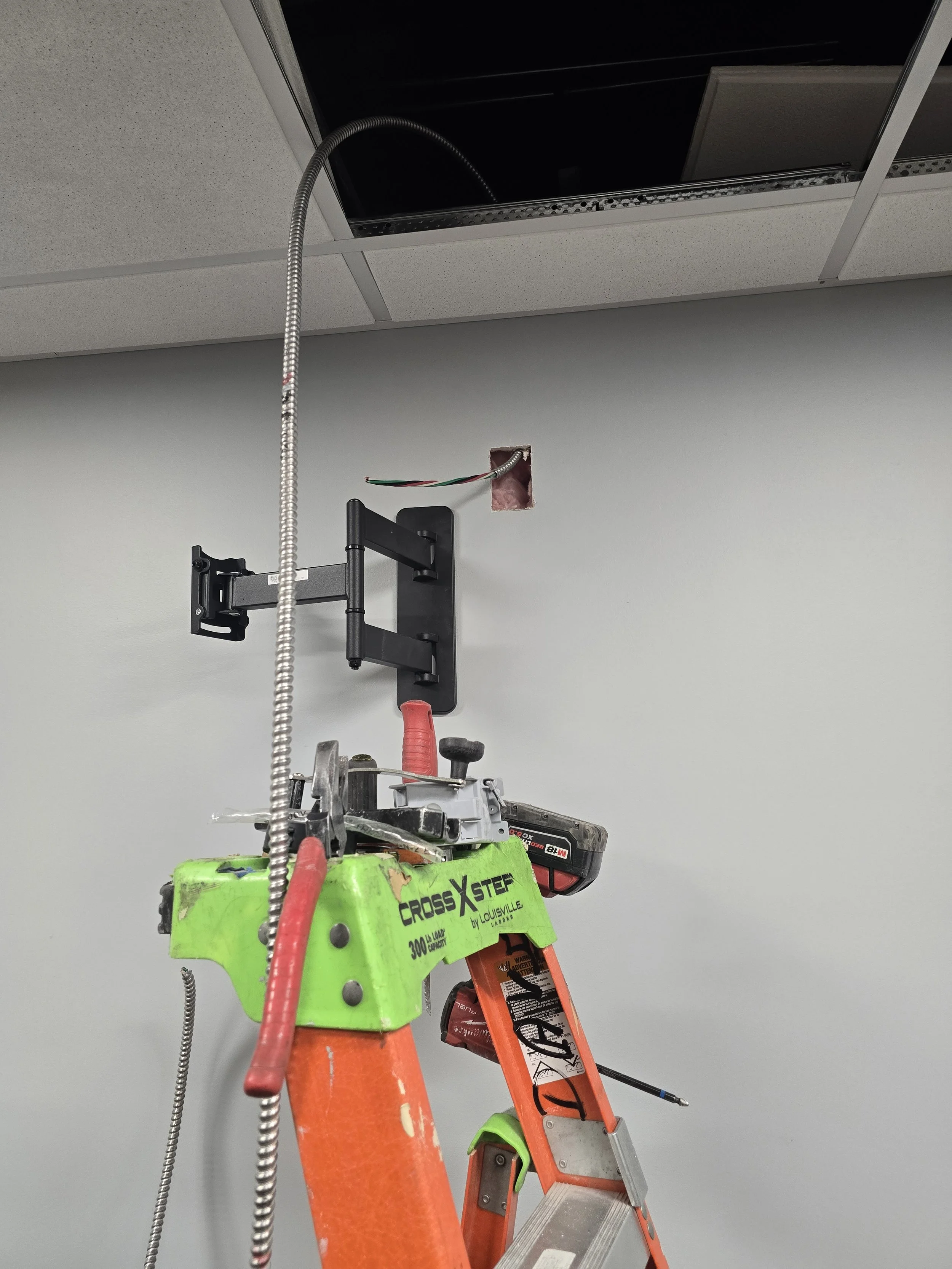 A portable work platform with an adjustable ladder, a work light, and electrical wiring extending toward a ceiling with an open ceiling tile, in an office or utility room setting.