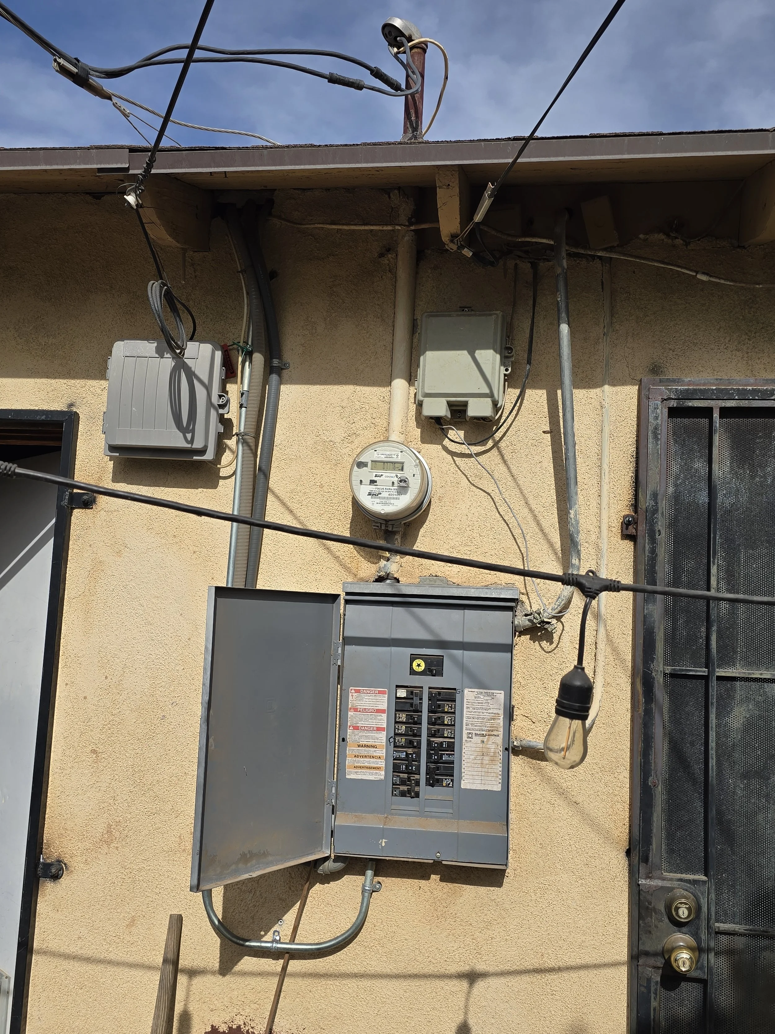 The image shows the exterior of a building with an electrical meter and fuse box mounted on the wall, along with various electrical wires and conduits.
