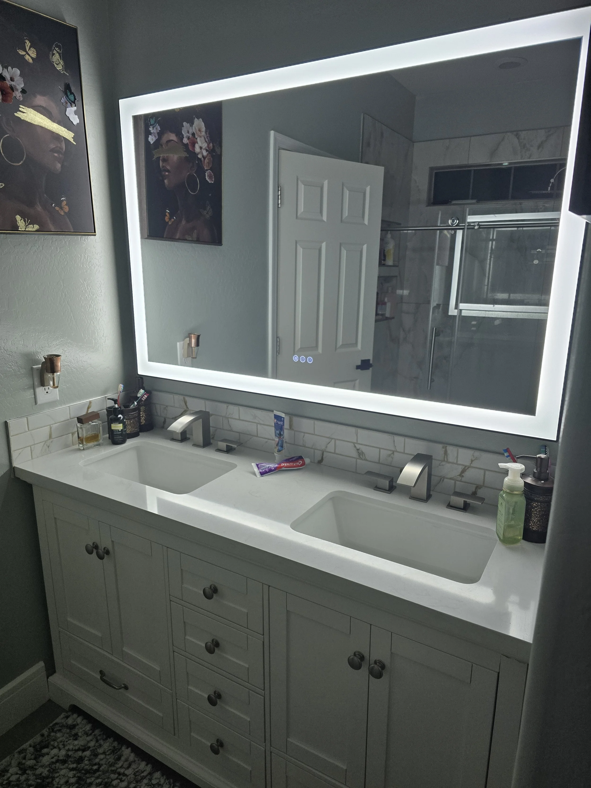 A bathroom vanity with a large illuminated mirror, a double sink, and various toiletries on the countertop. There is a shower with glass doors and a marble interior in the background.