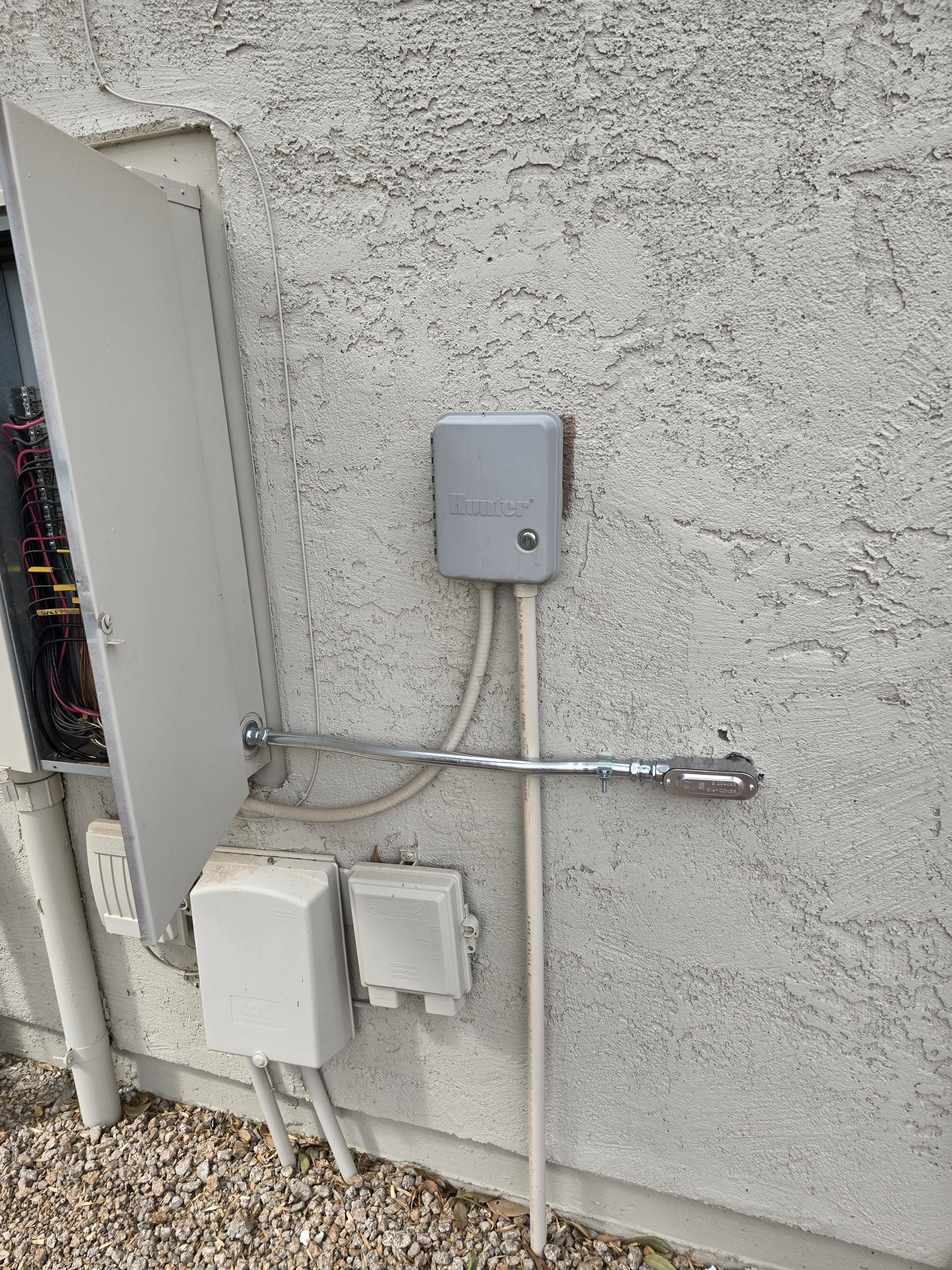 Close-up of outdoor electrical boxes and meters mounted on a textured white wall, with various wiring and conduit pipes connected to them, and gravel on the ground.