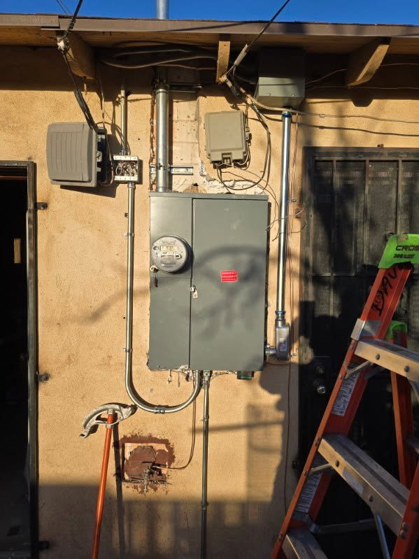 Electrical meter and panel box mounted on an exterior wall, with various electrical conduits and boxes, a ladder, and a nearby window.