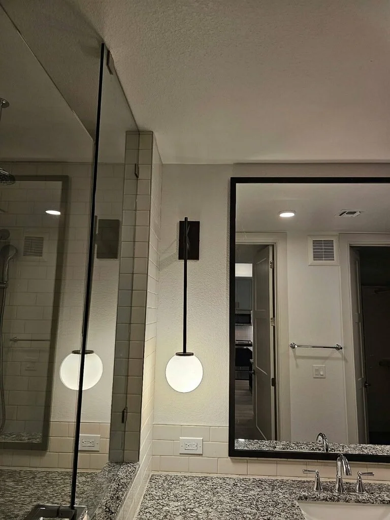 Bathroom with a large mirror, white tiled walls, granite countertop, illuminated globe pendant lights, and a walk-in shower with glass enclosure.