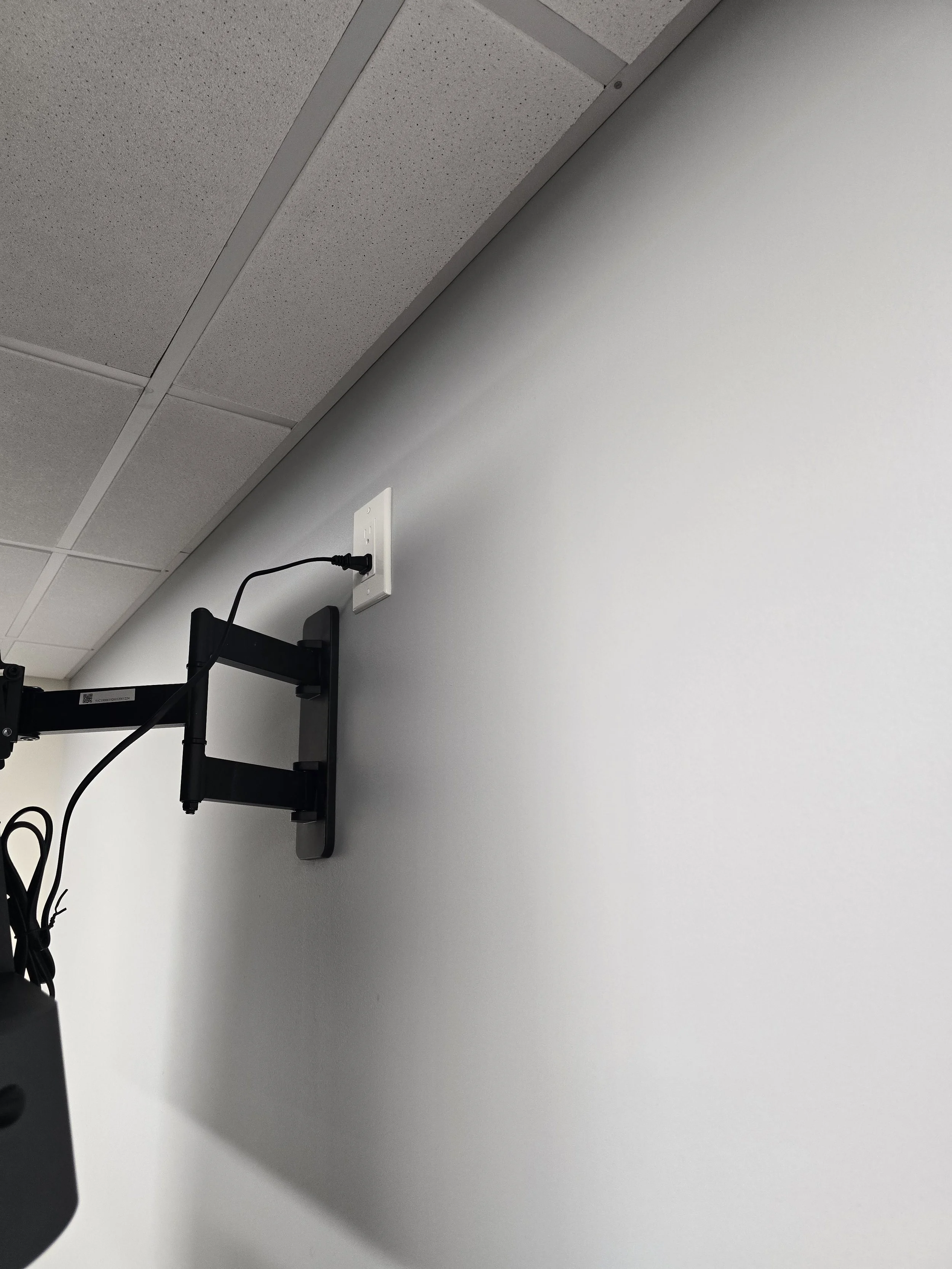 Ceiling tiles, wall with electrical outlet, and a monitor arm with a cable in an office or conference room.