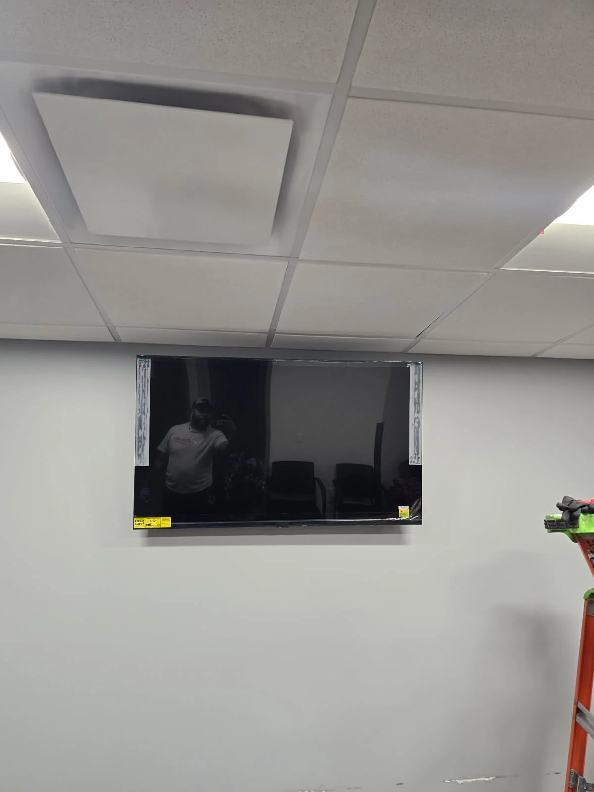 A wall-mounted flat-screen TV in a room with grey walls and a ceiling with square acoustic tiles. The TV is off, showing a reflection of a person taking the photo and some chairs in the background. Part of a stepladder and some equipment are visible 