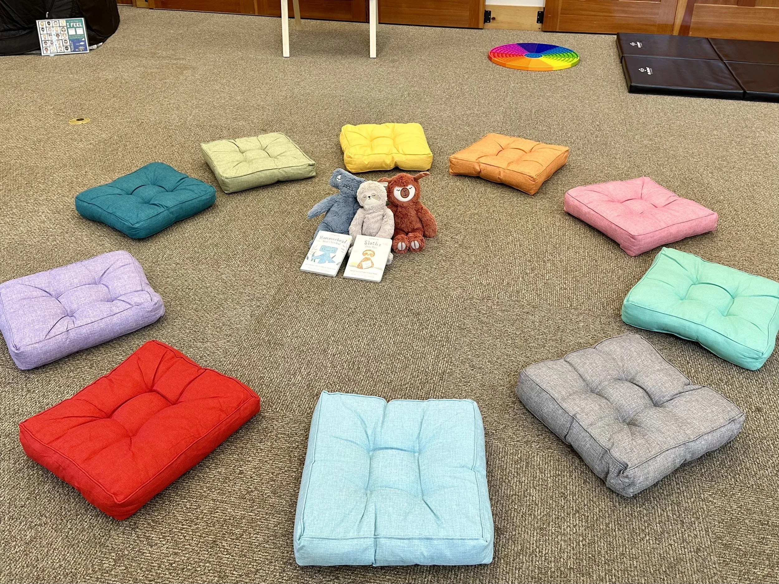 Colorful cushions arranged in a circle on a carpeted floor of a room, with plush animal toys, books, and various educational materials in the background.