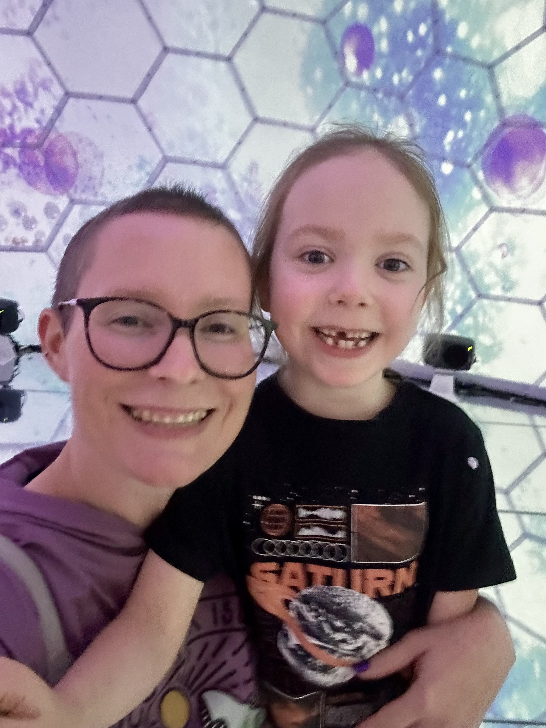 A woman and a young boy are taking a selfie together, smiling at the camera. The background has a colorful, patterned ceiling with hexagonal shapes.