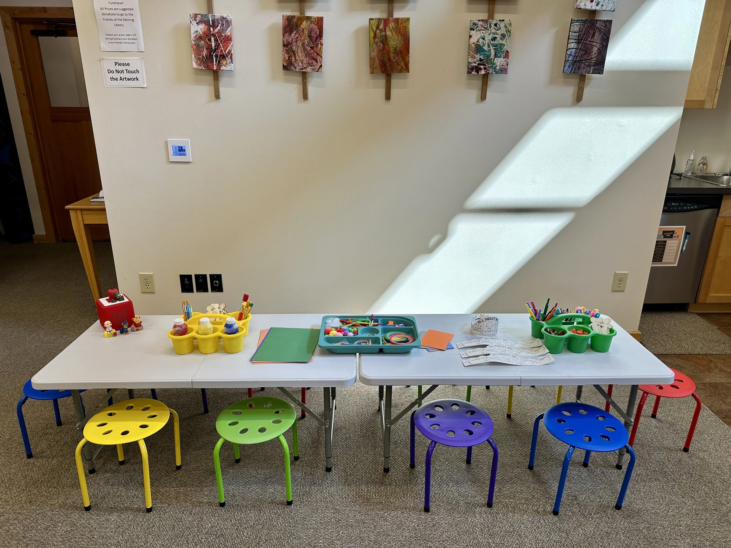 A craft table with colorful plastic stools around it, holding art supplies such as paper, markers, and various craft items. The background features a wall with abstract artwork pieces hanging and signs, with a small kitchenette area on the right.