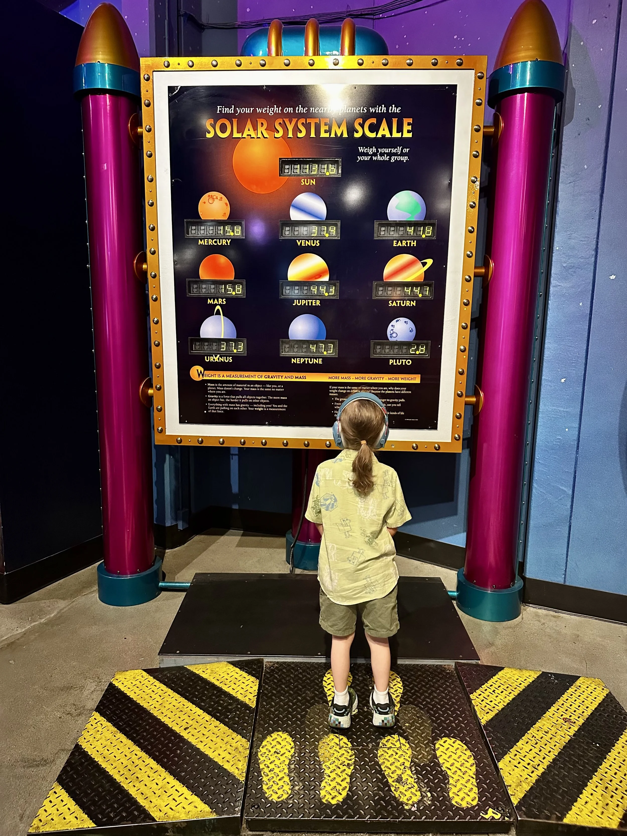 A young boy with a ponytail, wearing a yellow shirt and shorts, stands on a black and yellow striped scale in front of a colorful display about the solar system at a science museum. The display shows the sizes of planets relative to the sun.
