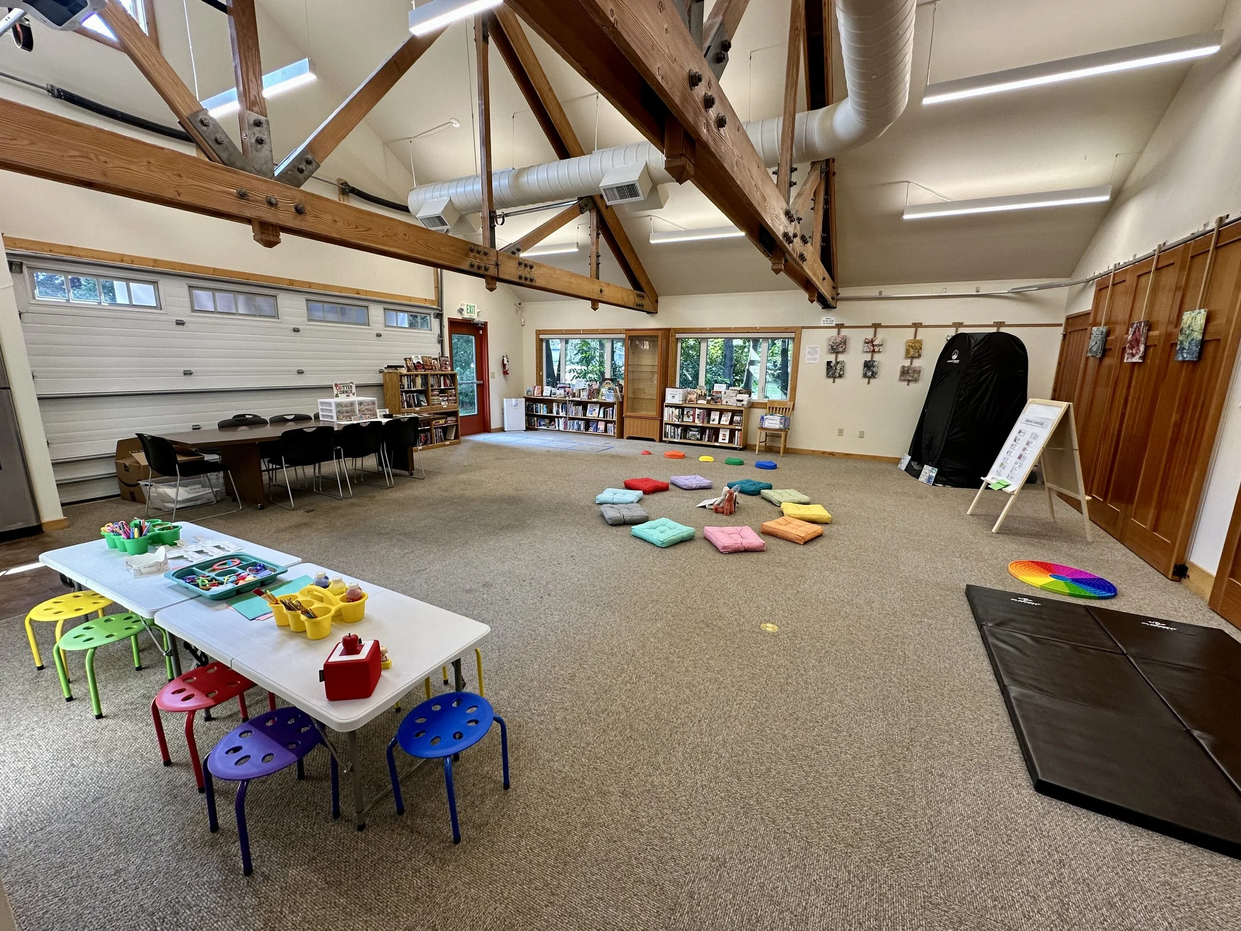 A spacious classroom or community center room with high ceilings featuring wooden beams and large windows. The room contains children's play and activity areas with colorful cushions, a small table with chairs, and various educational materials and books. There are shelves stocked with books, artwork on the walls, and play equipment, including a black mat and a small indoor climbing wall.