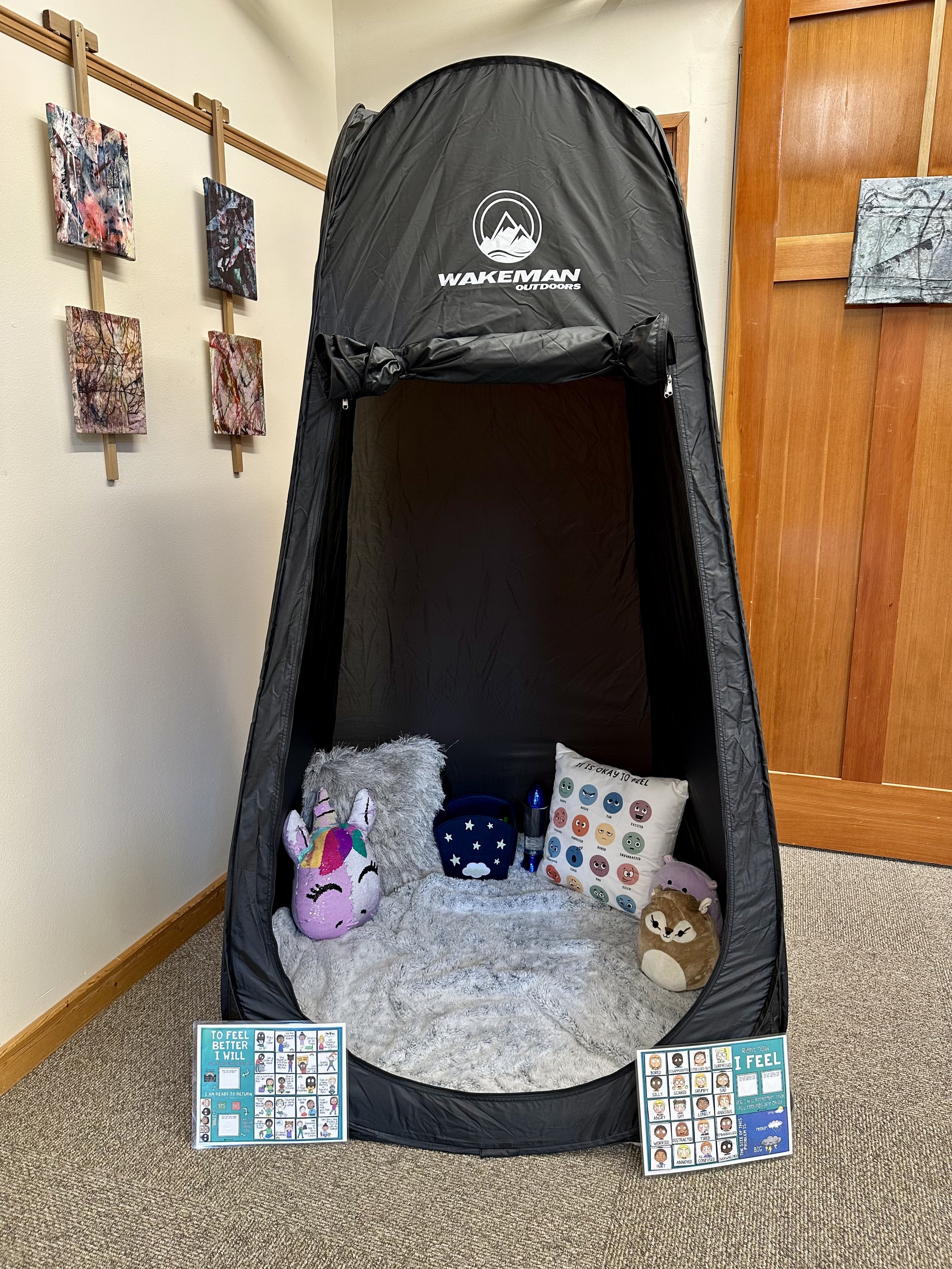 A sensory tent set up indoors with a plush gray blanket on the floor, surrounded by plush toys and mood emotion cards. Signs in front encourage emotional awareness and regulation, indicating it's for helping children feel better.