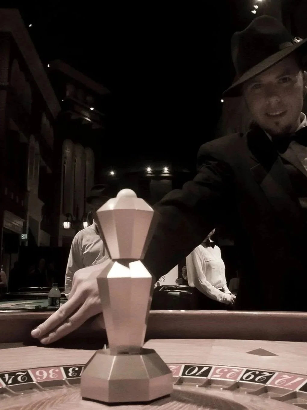 Close-up of a roulette wheel at a casino table with a prominent white chip in the foreground. A person wearing a gray fedora, tuxedo, and headphones is reaching towards the wheel, smiling. Other people and the interior of the casino are visible in the background.