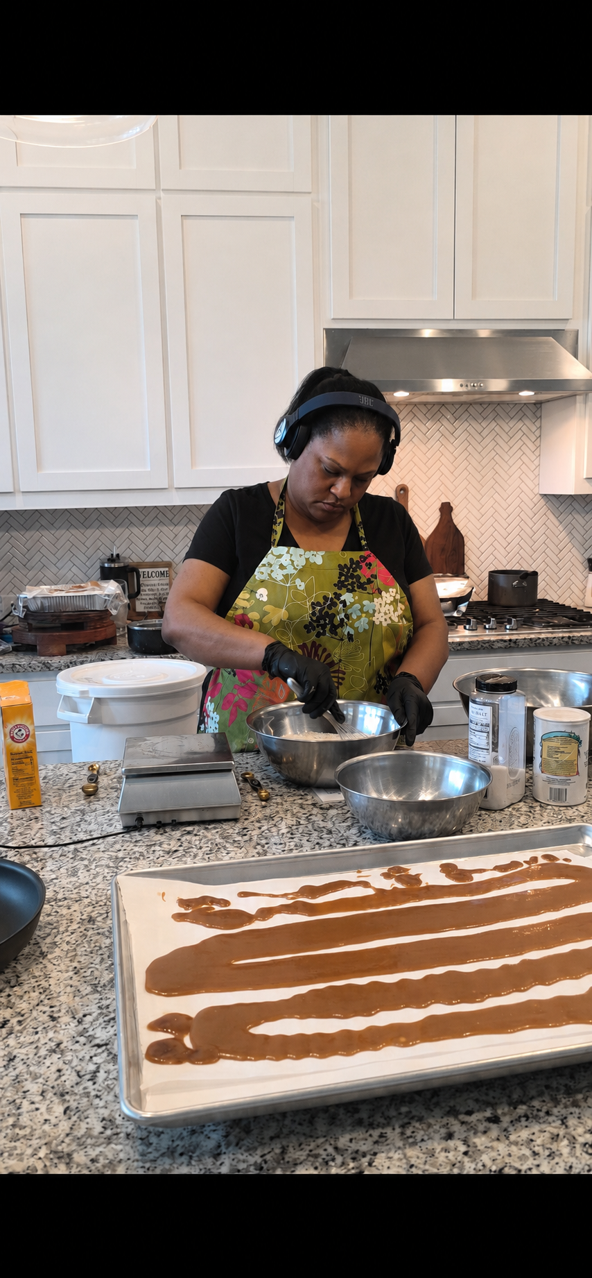 A woman in a colorful apron and black gloves is preparing chocolate in a mixing bowl in a modern kitchen, with a tray of raw chocolate at the foreground.