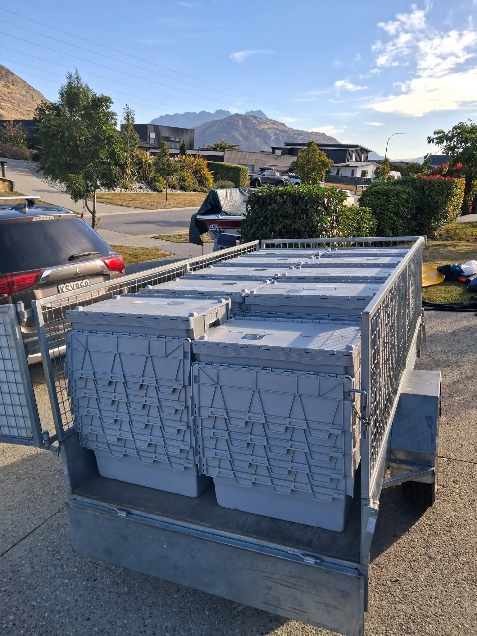 A fenced trailer filled with stackable moving boxes parked on a paved surface with some bushes and trees nearby, with mountains and houses in the background under a blue sky with some clouds.