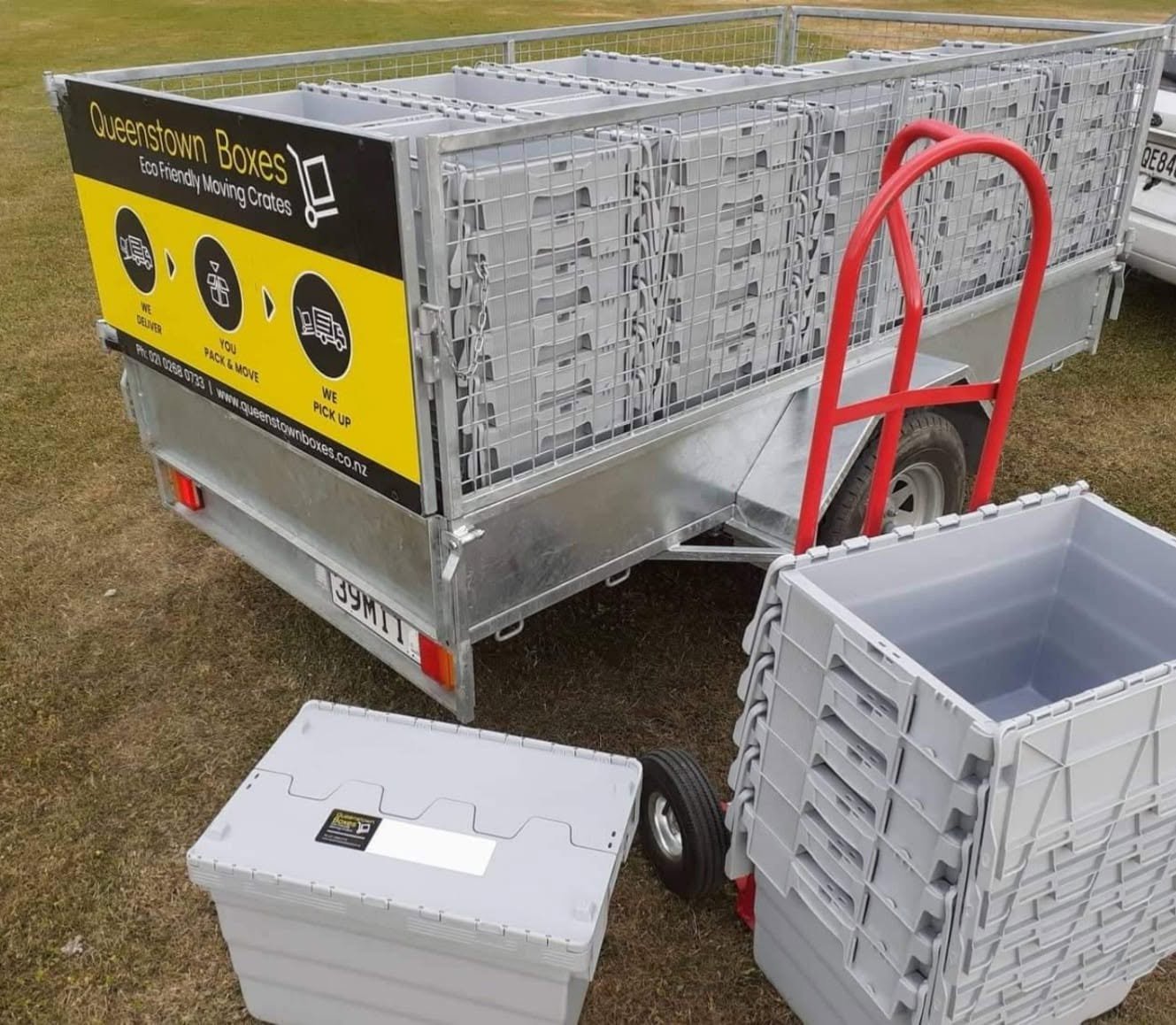 A trailer loaded with multiple gray plastic moving crates and a yellow and black sign that reads 'Queenstown Boxes, Eco Friendly Moving Crates.' There is a red hand truck leaning against the trailer, and additional empty crates are placed nearby on the ground.