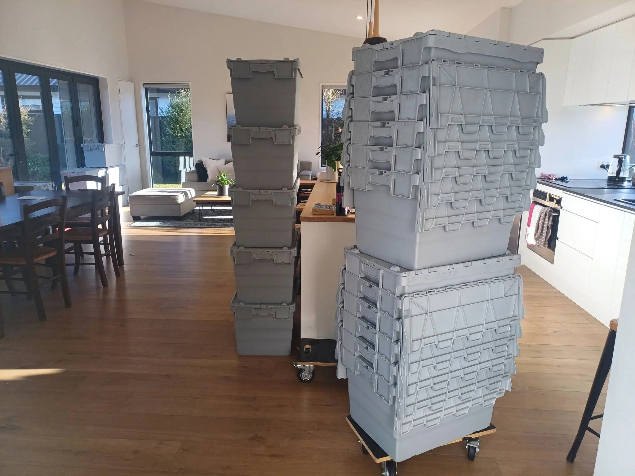 Stack of plastic moving boxes on a wheeled cart in a bright room with large windows, dining table, and living area.