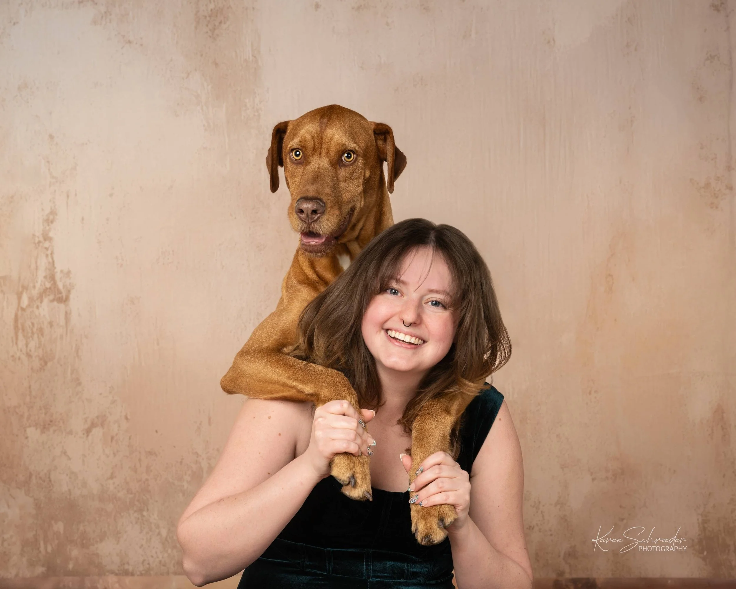 A woman with light skin and shoulder-length brown hair smiling while holding a brown dog with golden eyes on her shoulders. The background is a textured beige wall.