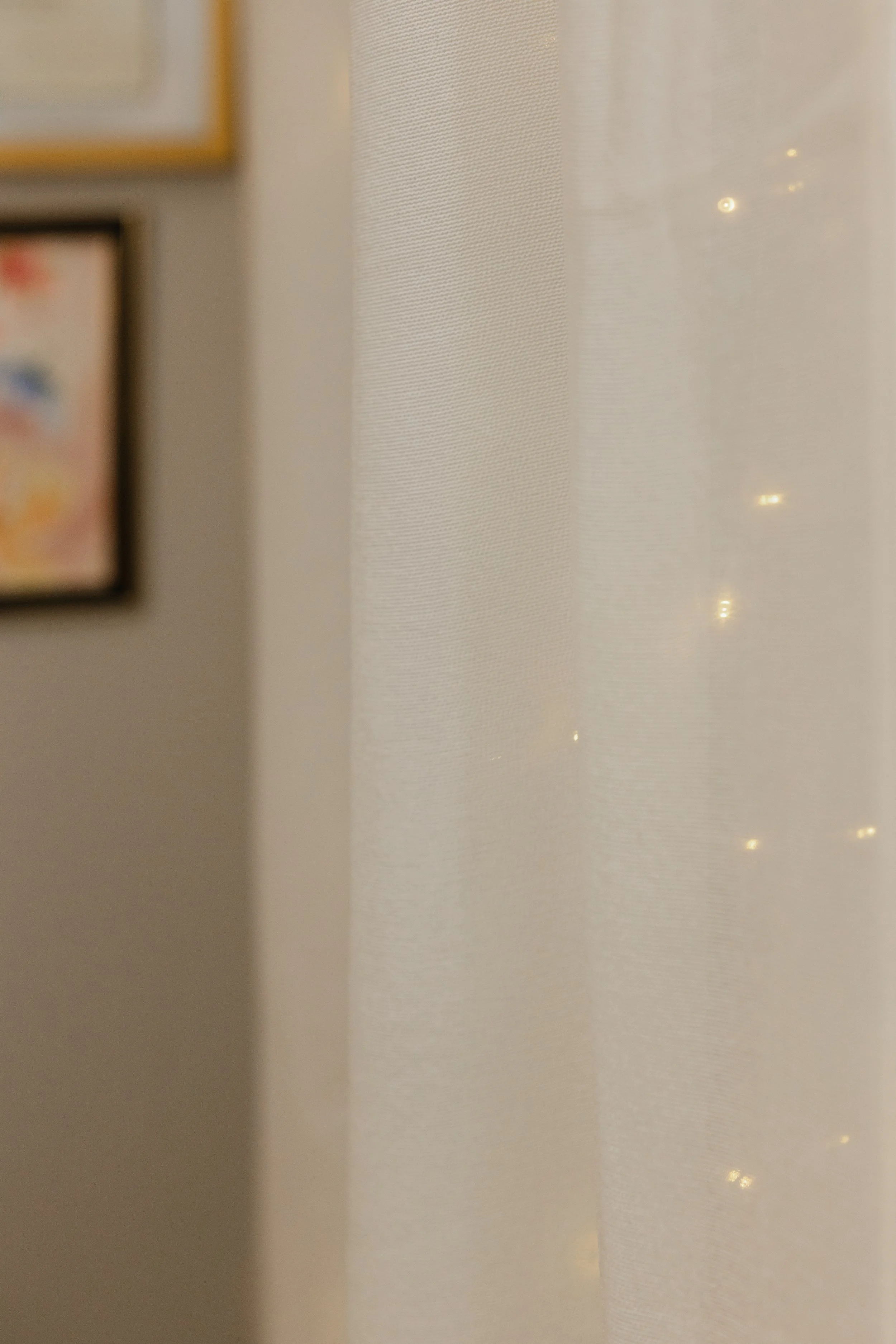 Partially visible framed artwork on wall next to a sheer curtain with small string lights behind it.