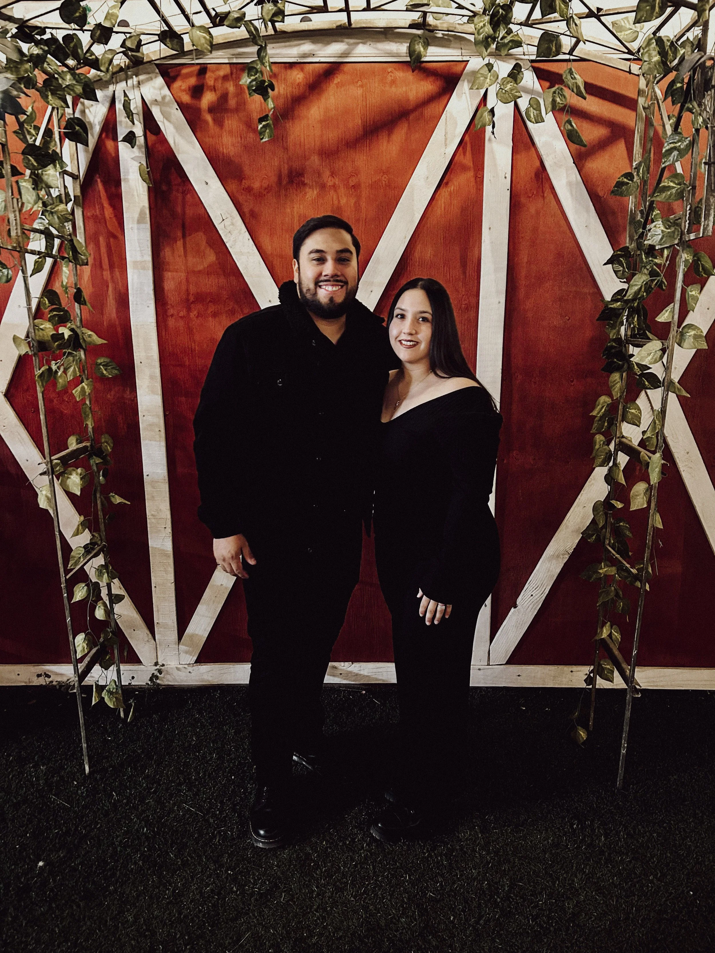 A man and woman standing together in front of a red barn-style wall decorated with white trim and outdoor string lights. The man has a beard and is wearing a black jacket, and the woman has long dark hair and is wearing a black off-the-shoulder top.