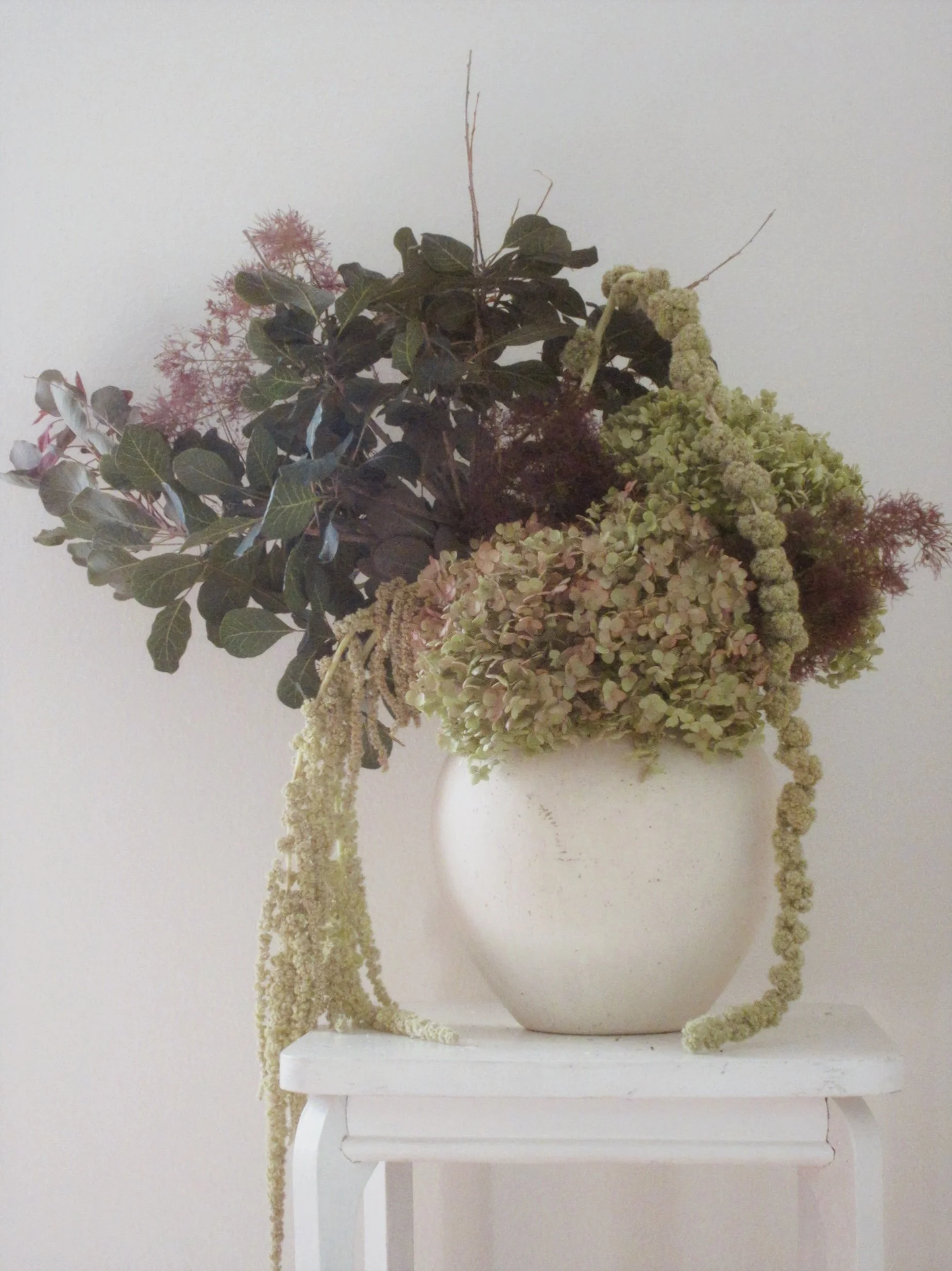White ceramic vase with dried flowers and plants, such as hydrangeas and possibly eucalyptus, placed on a white table against a plain white wall.