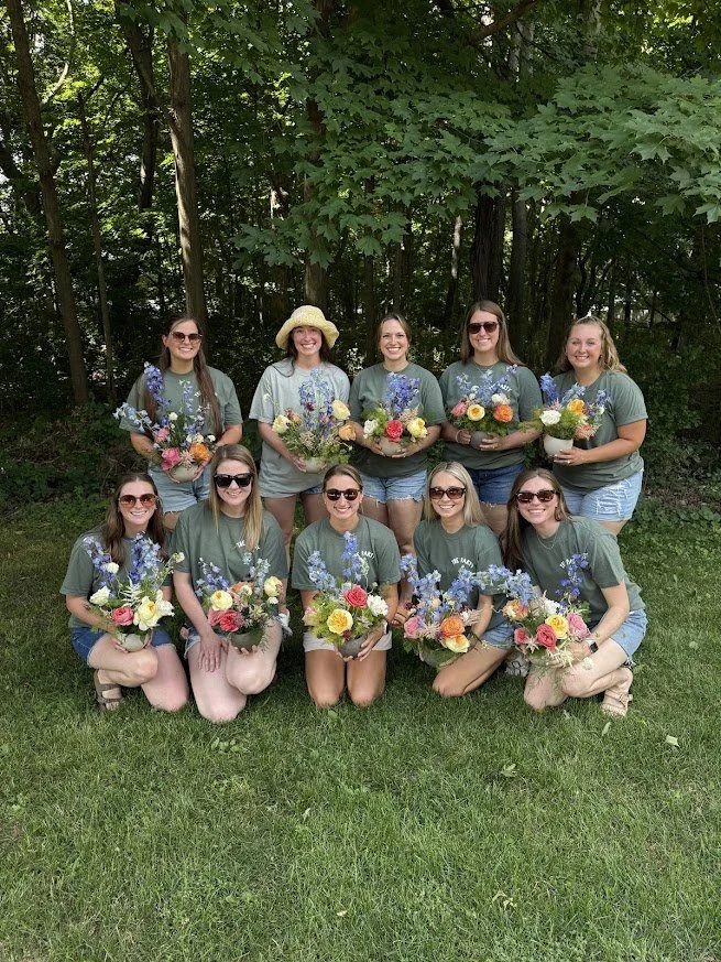 A group of ten women outdoors in a forested area, holding colorful flower bouquets, posing for a photo.