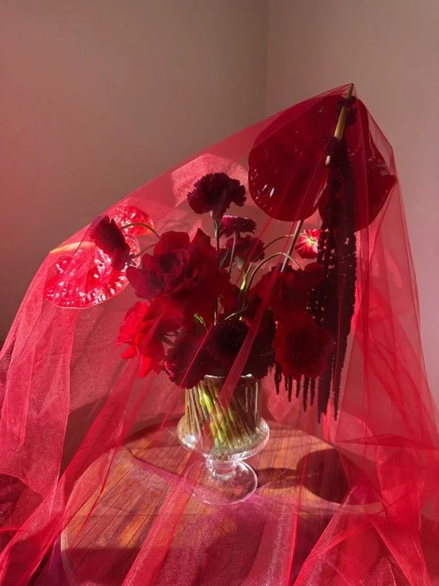 Red flowers arranged in a glass vase, covered by red sheer fabric and parasols, placed on a wooden table against a plain wall.