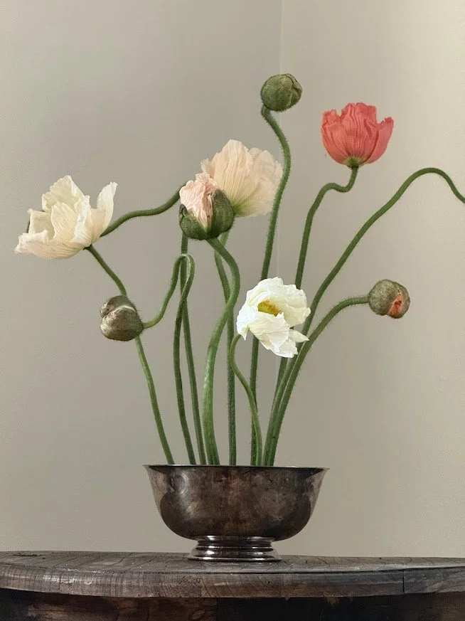 A metallic bowl holding a bouquet of poppy flowers with green stems and large buds, placed on a wooden surface against a plain wall.